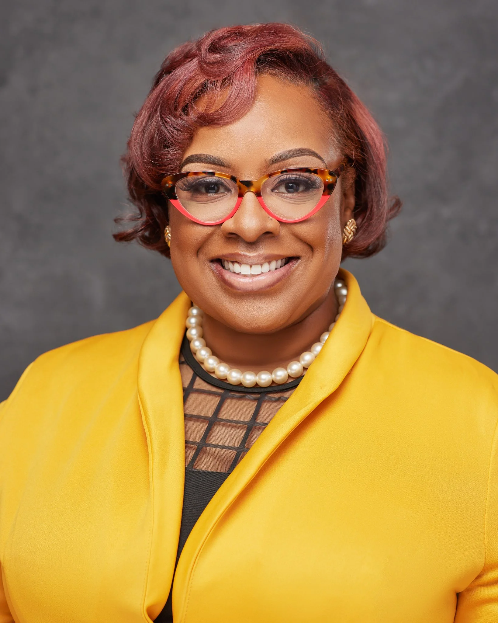 Portrait of a woman with short red hair, wearing glasses with red and tortoise shell frames, a pearl necklace, gold earrings, a black top with a checkerboard lace pattern, and a yellow blazer, smiling in front of a gray background.