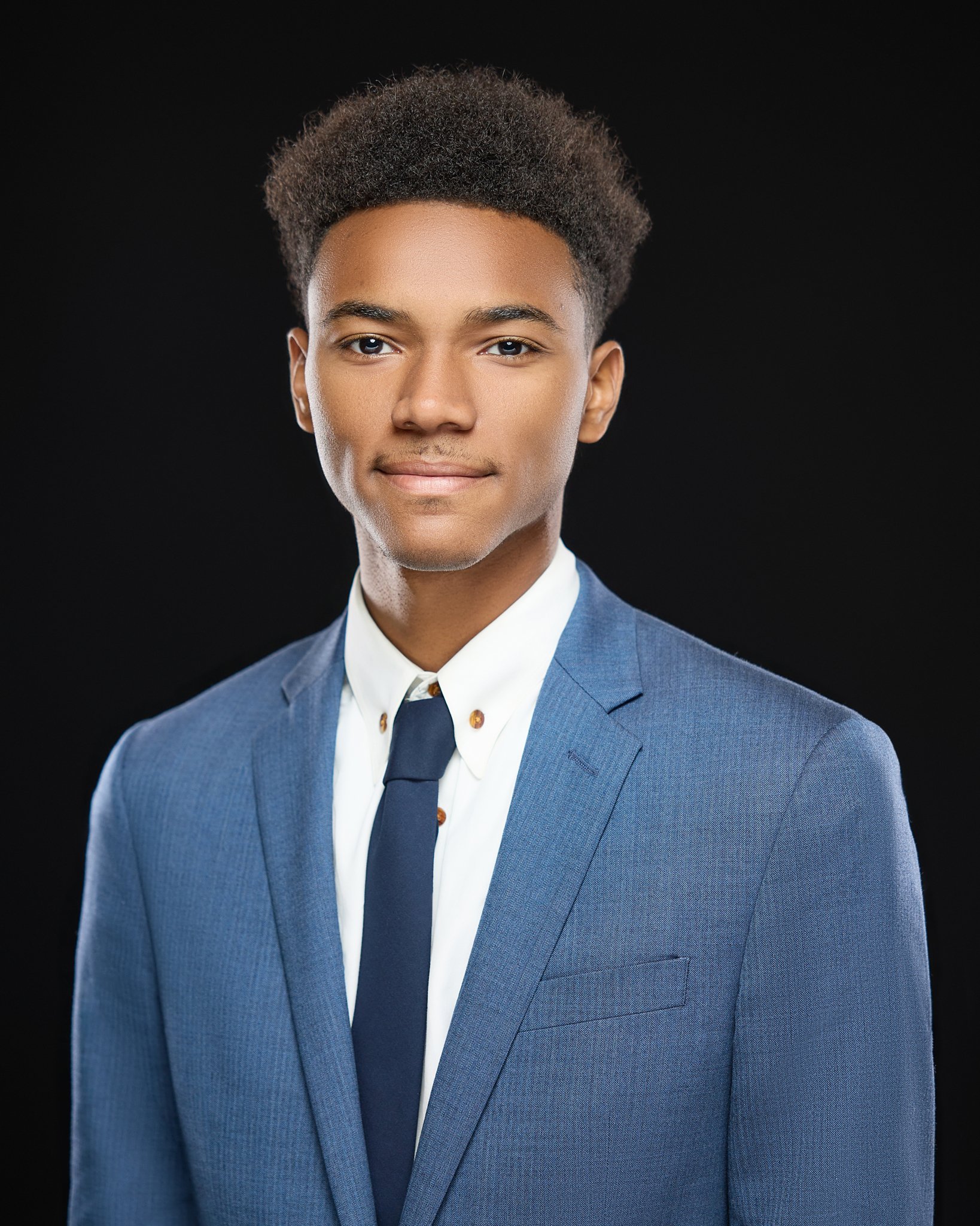 Young man in a blue suit, white shirt, and dark tie, smiling against a black background.
