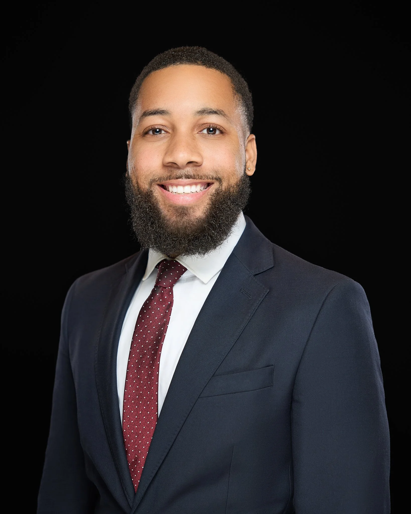 A professional Headshot of a young man with a beard and short hair, smiling, wearing a dark suit, white shirt, and maroon tie with white dots, against a black background.