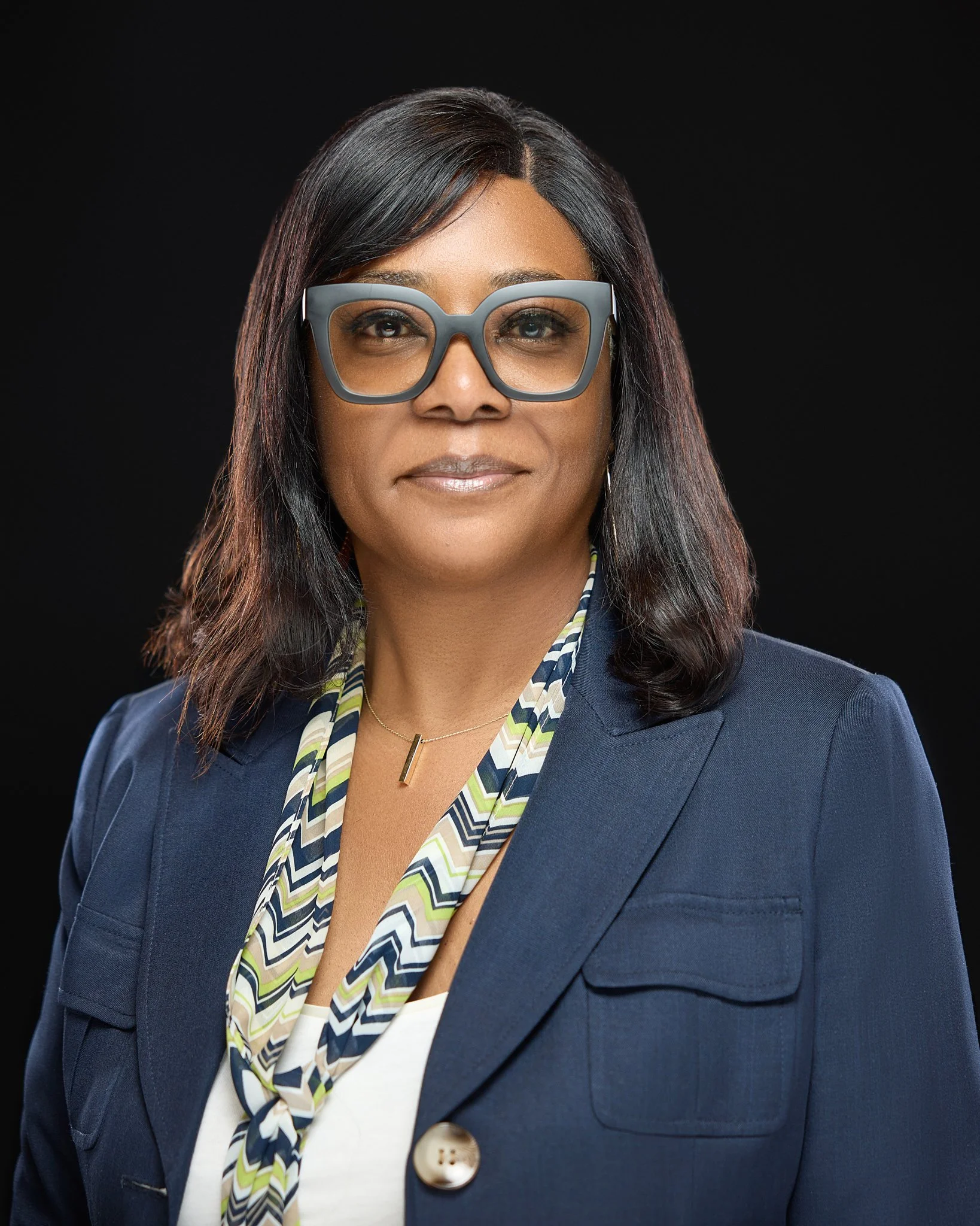 A professional Headshot of a woman with dark hair, glasses, and a navy blazer poses against a black background.