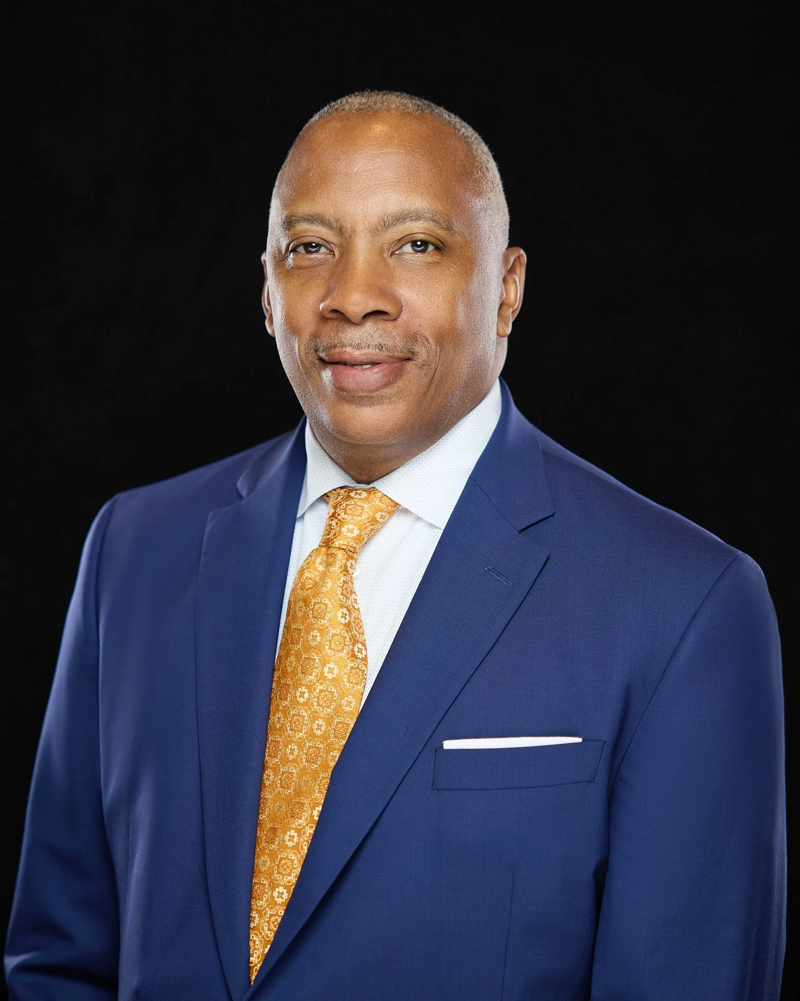 A headshot of a professionally dressed man in a blue suit, white shirt, and gold tie against a black background.