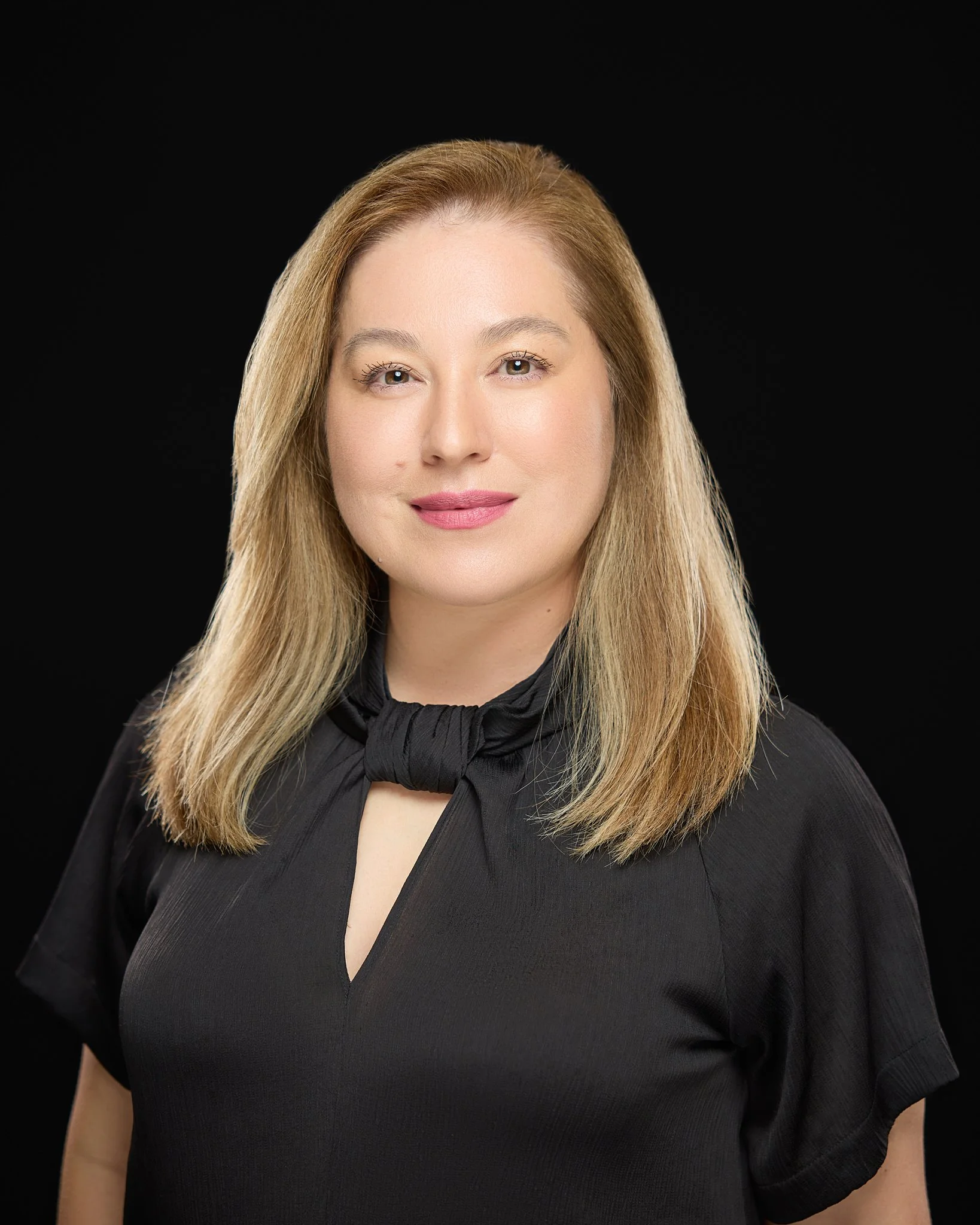 A professional headshot a woman with shoulder-length blond hair, light makeup, wearing a black top with a bow detail at the neckline, standing against a black background.