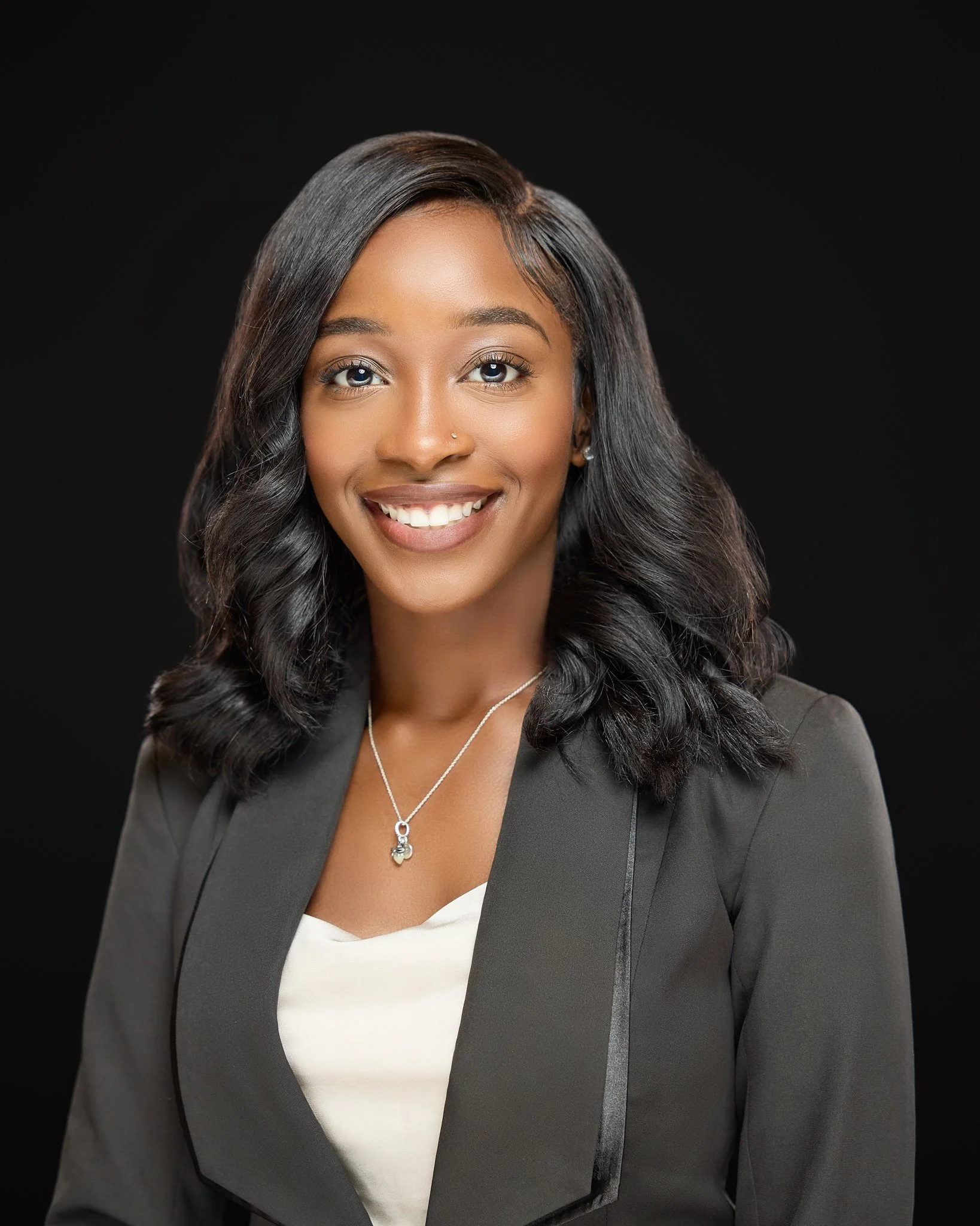 Professional headshot of a smiling young woman with dark hair, wearing a gray blazer, white top, and a necklace, against a black background.