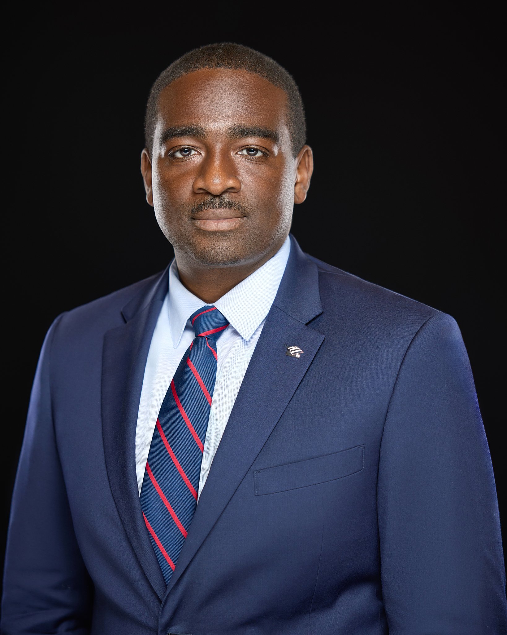 A professional headshot of a man in a navy suit, light blue dress shirt, and a striped red and blue tie, posing against a black background.