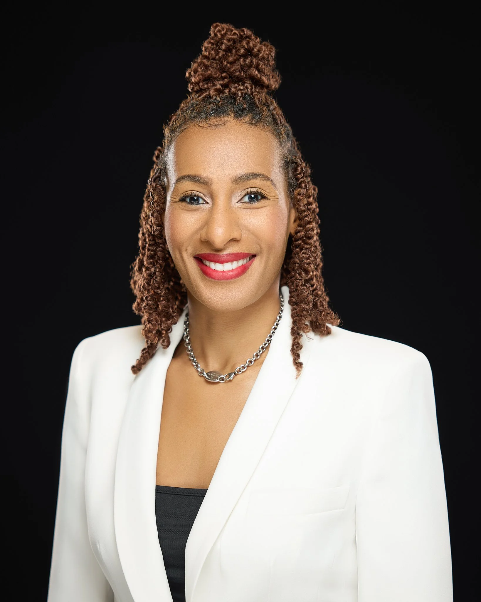 A woman with curly brown hair styled in a high bun and natural makeup, smiling, wearing a white blazer over a black top, accessorized with a silver chain necklace, against a black background.