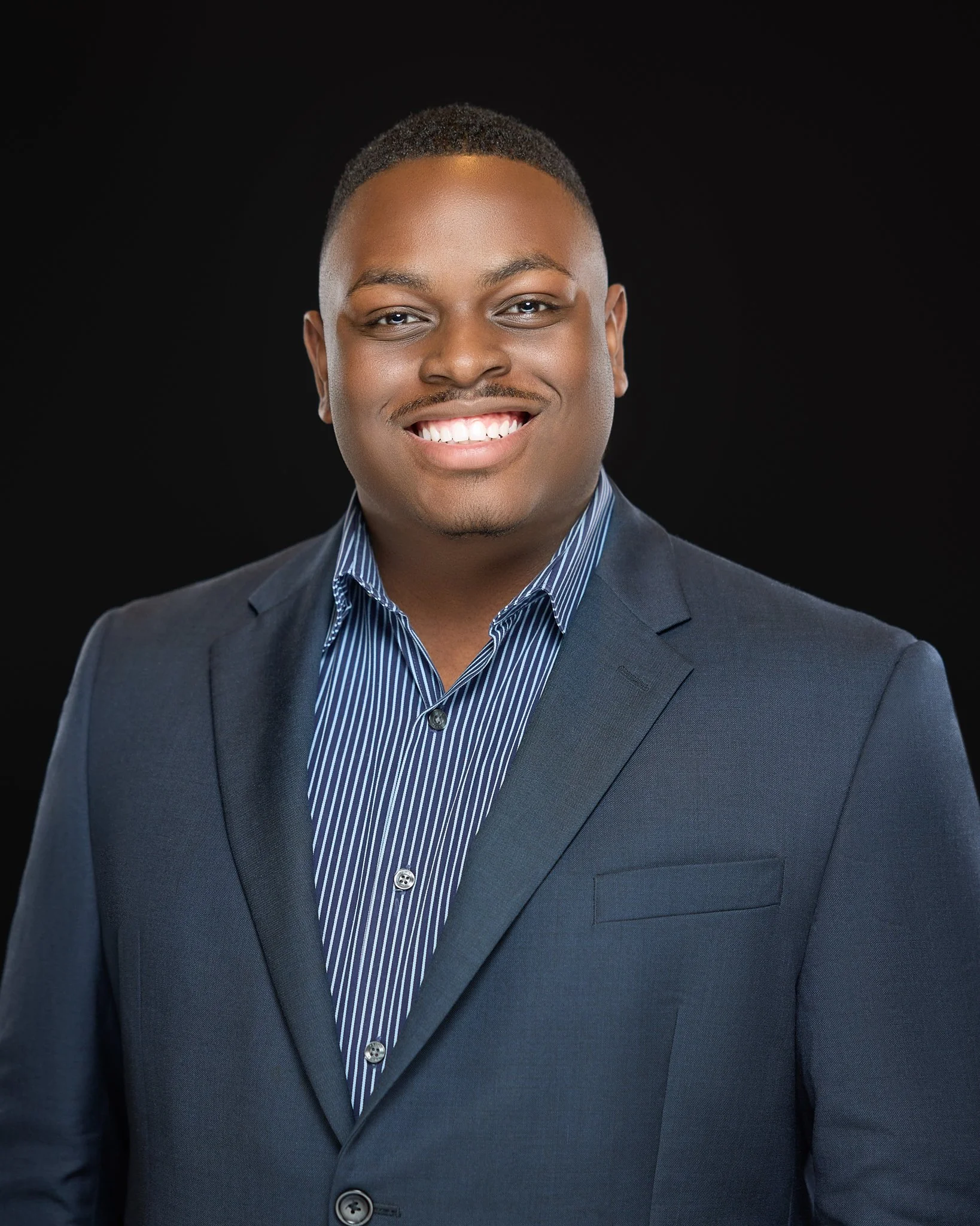 Portrait of a young man in a suit with a blue striped shirt, smiling against a black background.