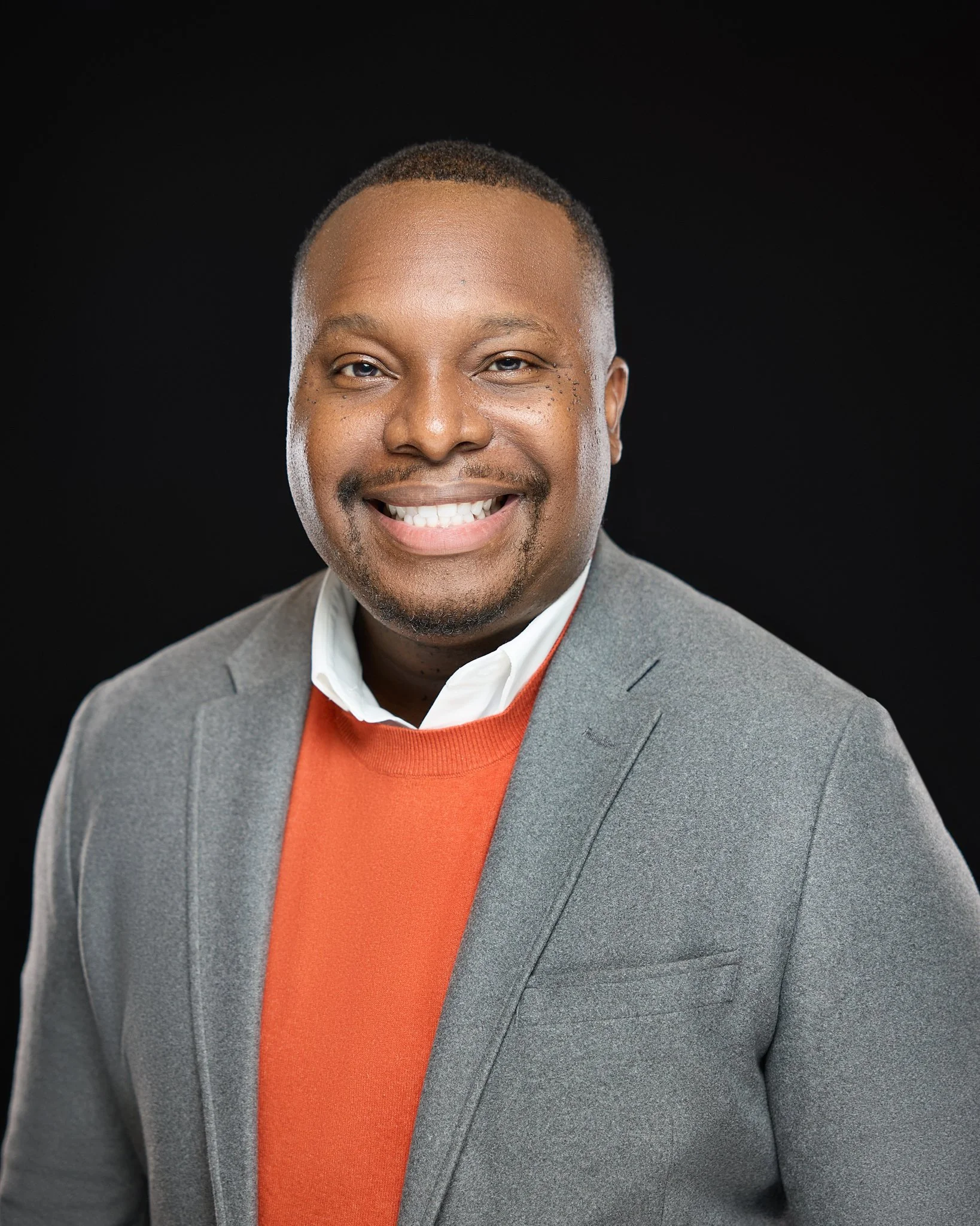 Professional portrait of an African American man smiling, dressed in a gray suit and orange sweater, against a black background.
