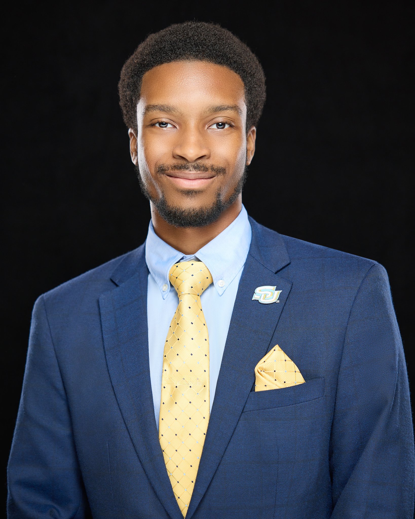 A young man with a confident smile, wearing a navy blue suit, light blue dress shirt, yellow polka dot tie, matching pocket square, and a university lapel pin, against a black background.
