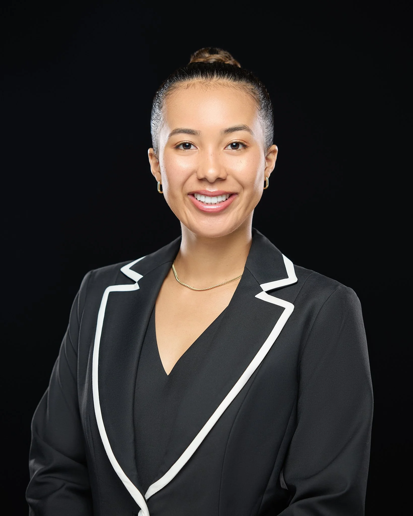 A professional headshot of a woman with a bun hairstyle smiling, wearing a black blazer with white trim, a black top, gold jewelry, and a black background.