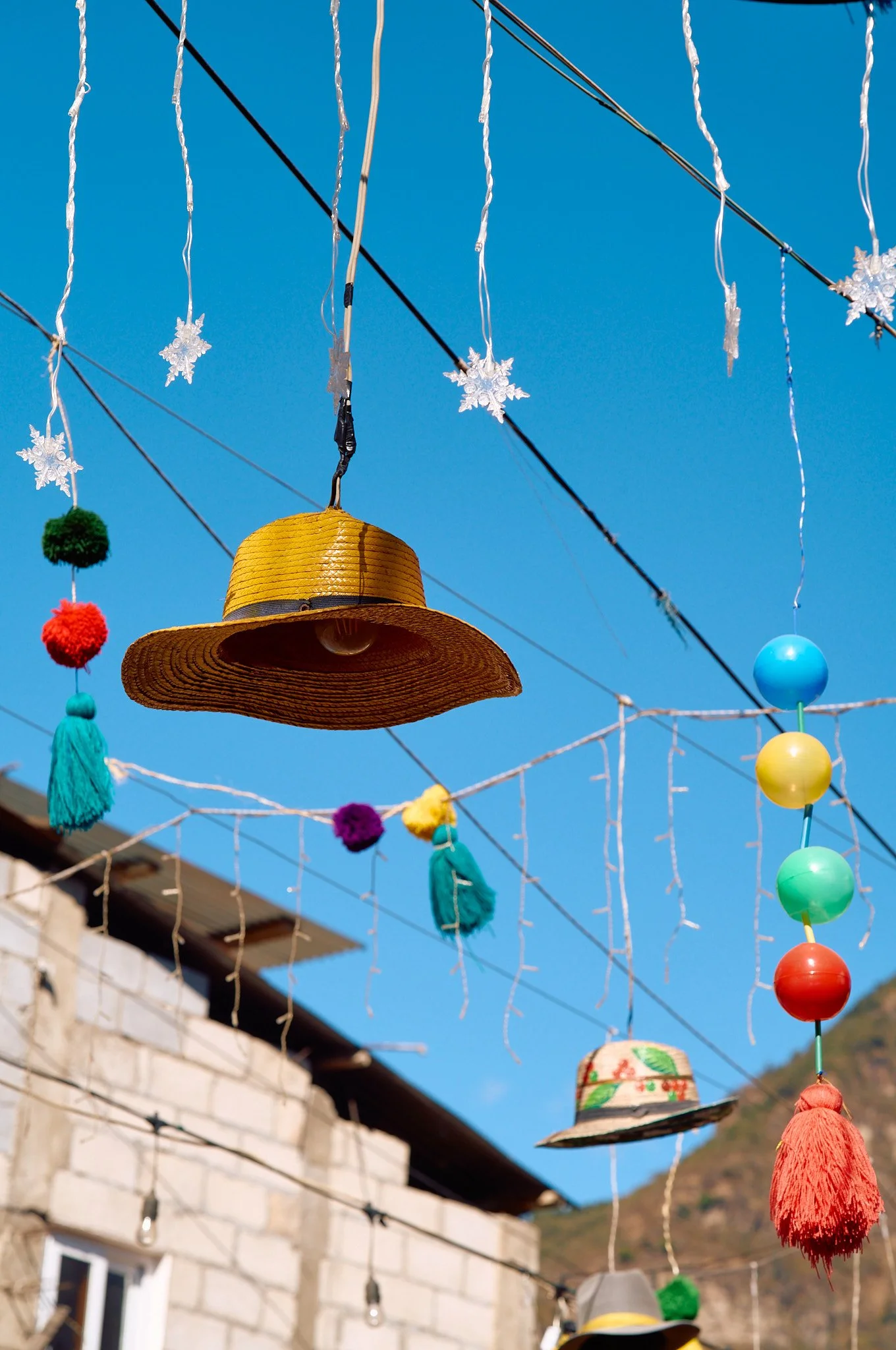 Colorful hanging decorations including hats, pom-poms, tassels, and snowflake ornaments hanging against a clear blue sky.