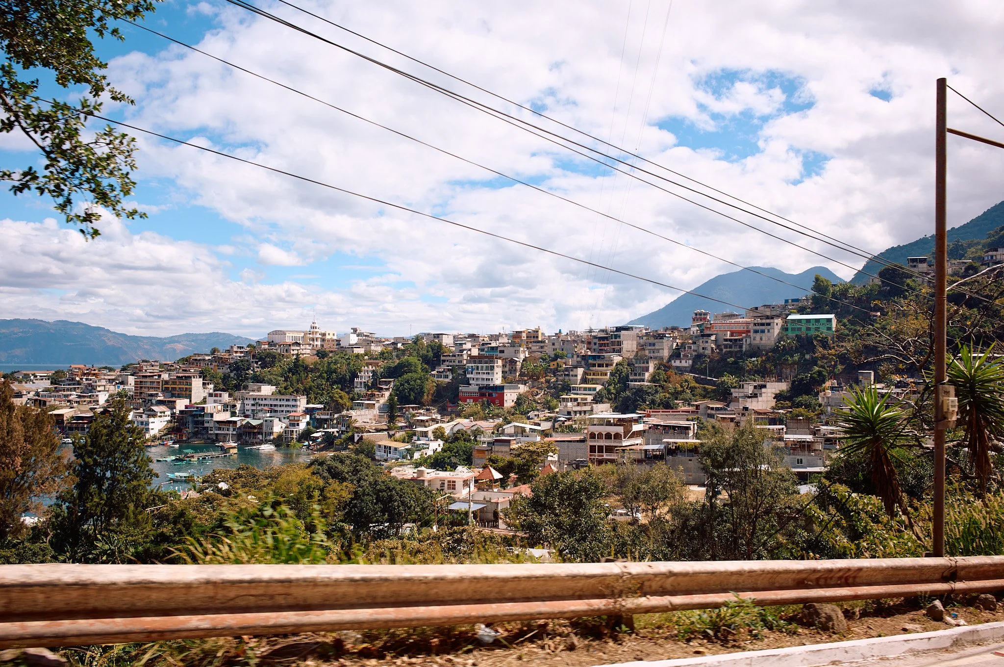 Hilly town with many multistory buildings on a hillside, surrounding a body of water, with mountains in the background and a partly cloudy sky.