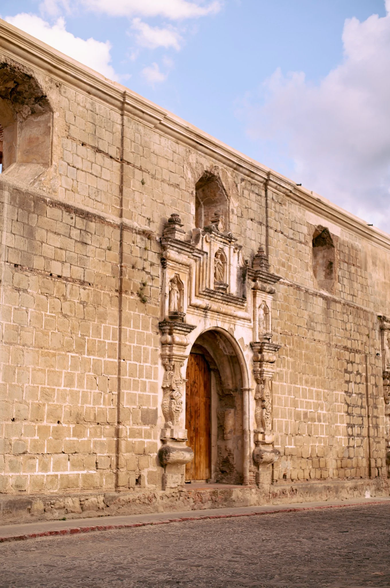 Stone building with ornate doorway and small niches, under a blue sky with clouds.