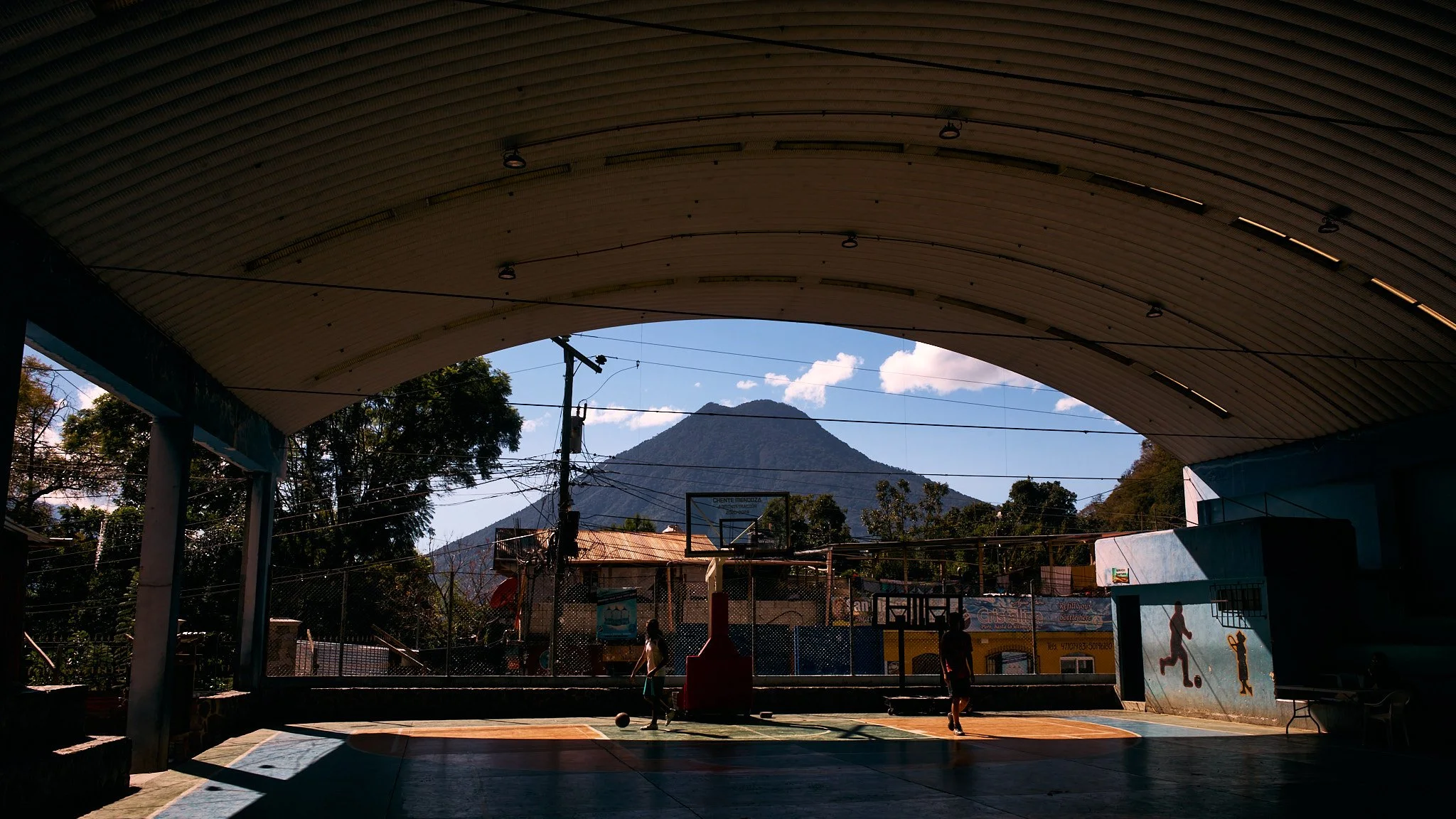 Under a large indoor basketball court roof, children are playing basketball and soccer, with a mountain in the background and power lines overhead.