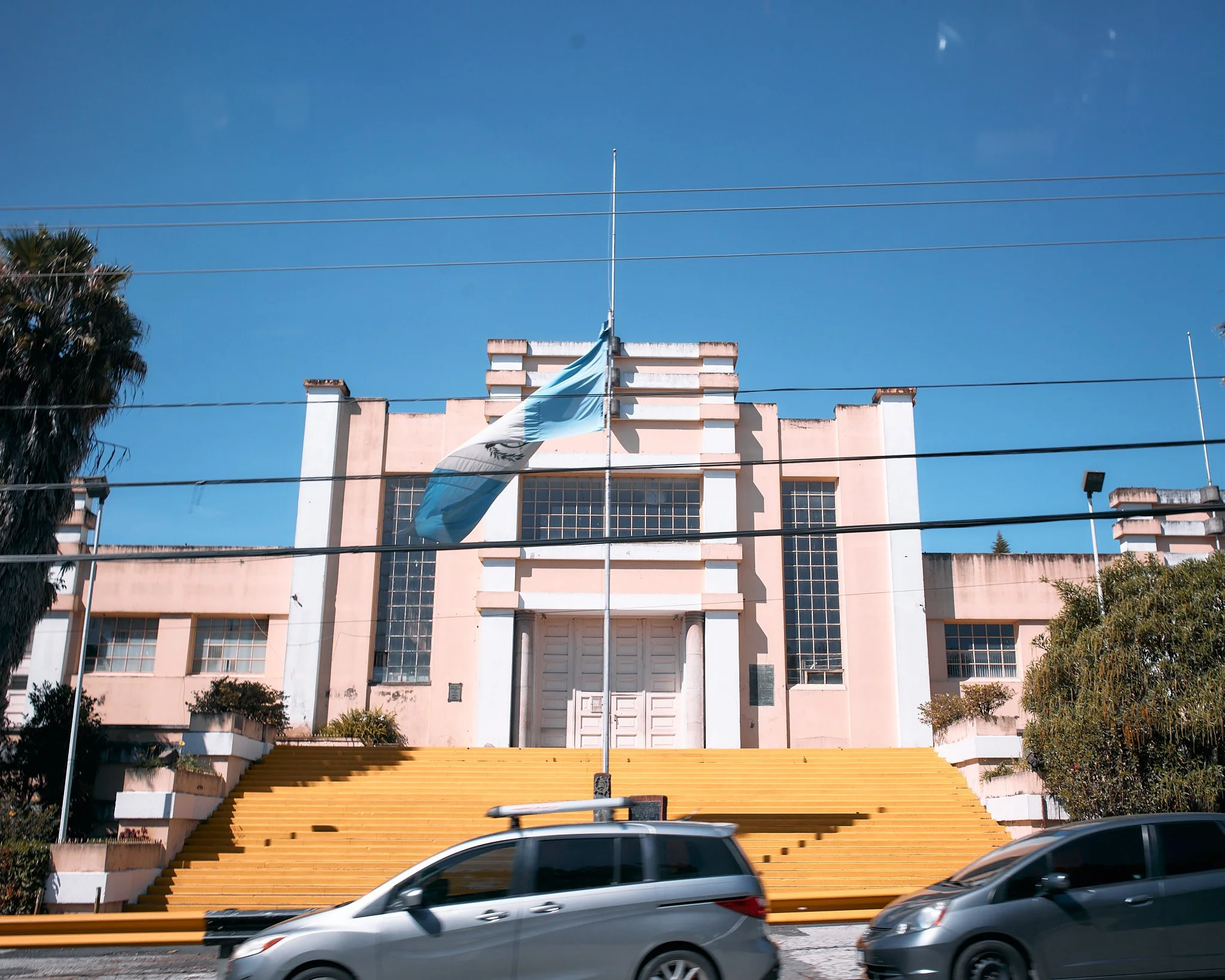 A large pink building with an Art Deco style, steps with yellow stairs leading up to the entrance, soy cars in front, and an Argentine flag flying in front.