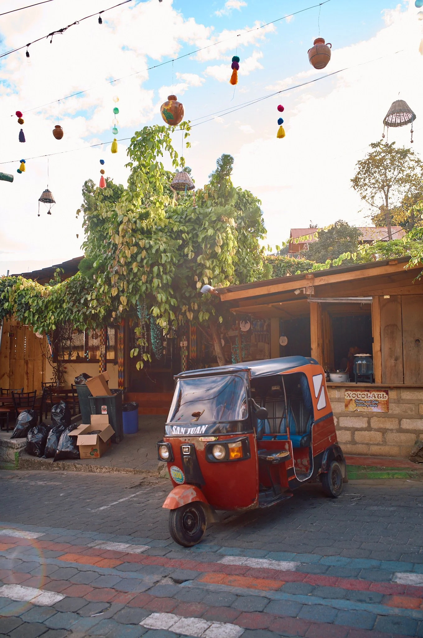 A red tricycle parked on a cobblestone street in front of a wooden building with a tree and hanging decorative lanterns and ornaments overhead.