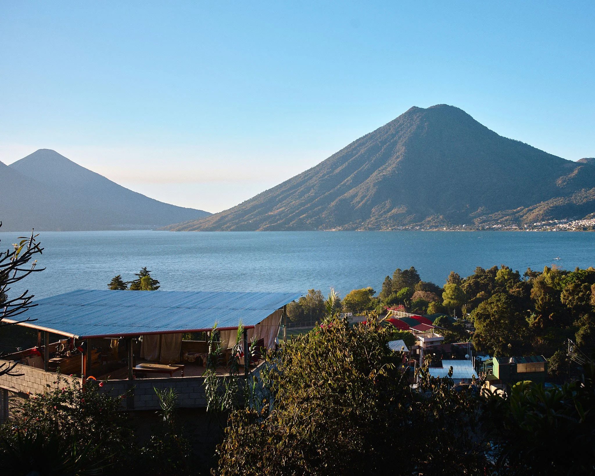 Volcanoes near a lake with houses and trees in the foreground and clear blue sky