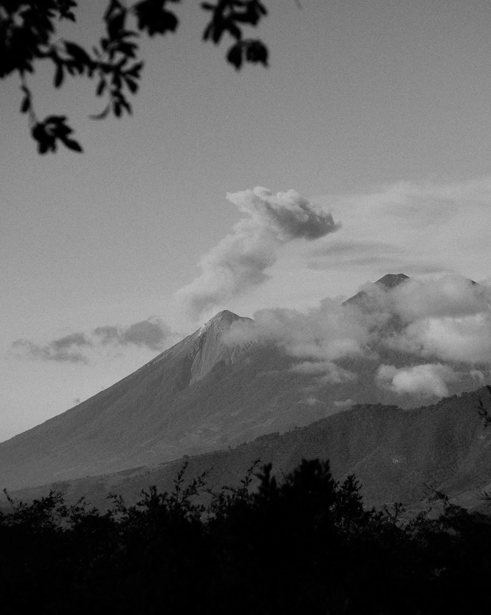 Black and white image of an erupting volcano with smoke and ash rising from the summit, surrounded by clouds and trees in the foreground.