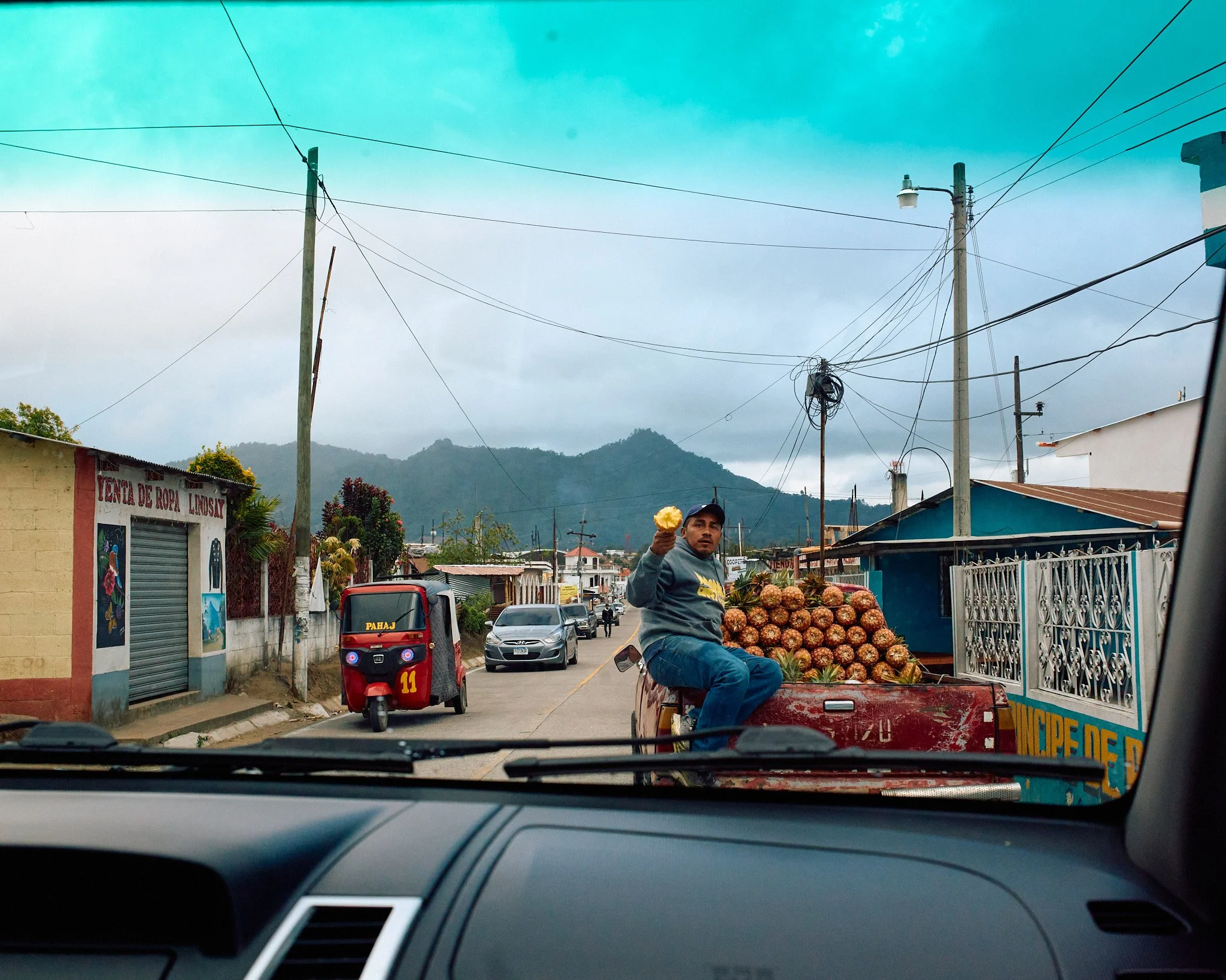 View from inside a vehicle showing a man selling pineapples on the back of a small truck in a street with mountains in the background. The man is holding an ice cream, sitting on the truck, which is parked among other vehicles.