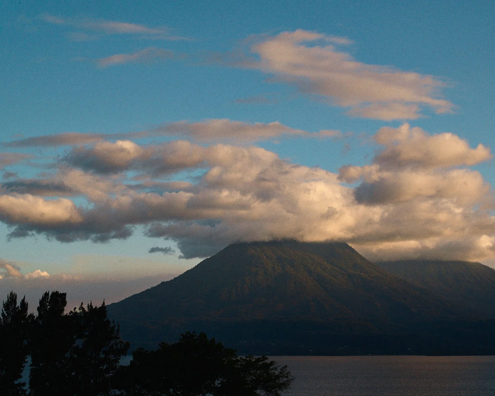 A mountain with a cloud formation above it, a body of water in the foreground, and trees at the bottom.