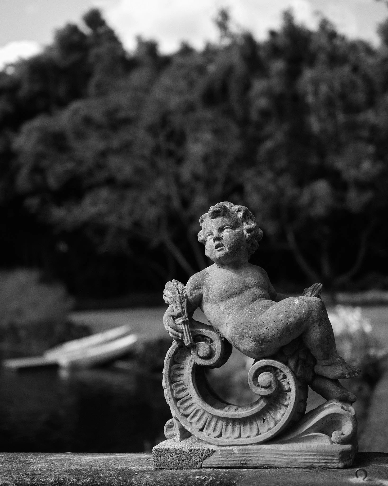 Black and white photo of a cherub sculpture sitting on a decorated scroll, outdoors with bushes and trees in the background.