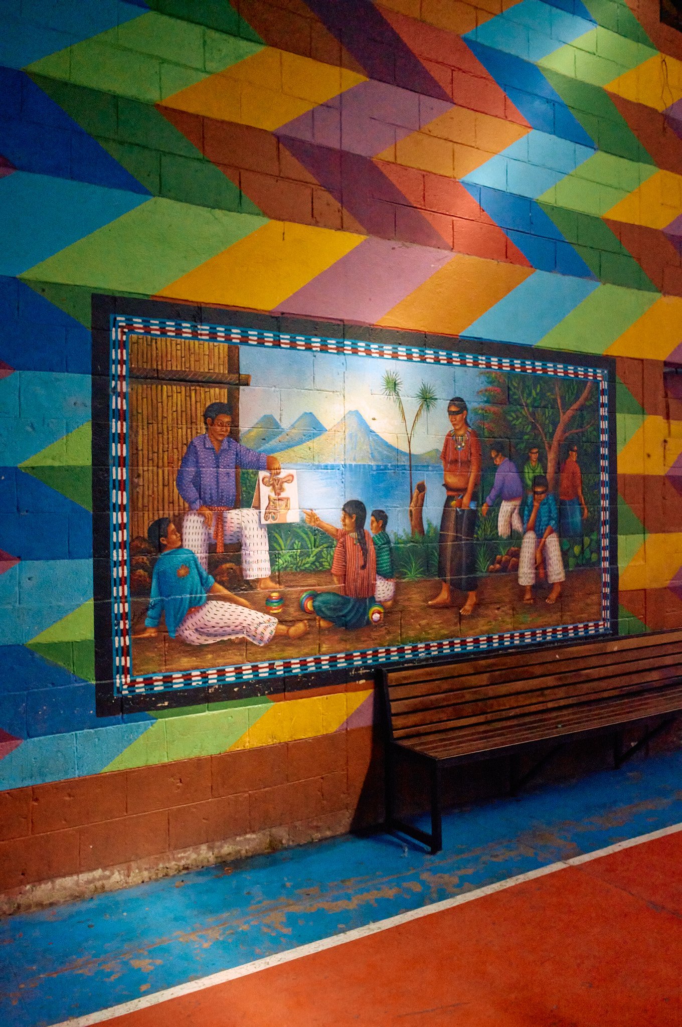 Colorful mural depicting indigenous people performing a traditional activity outdoors with mountains and trees in the background, framed by a vibrant geometric painted wall. A wooden bench is in front of the mural.
