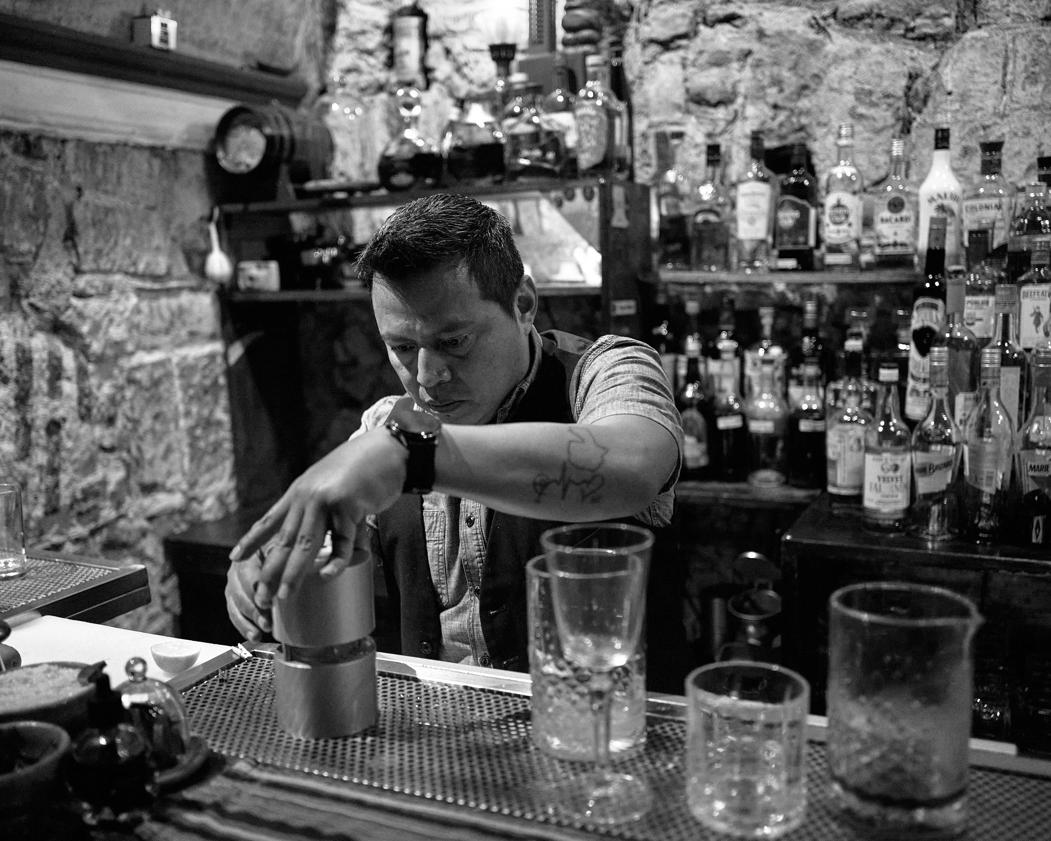 A bartender preparing a drink at a bar with a stone wall background, various liquor bottles on shelves, and several glasses on the counter.