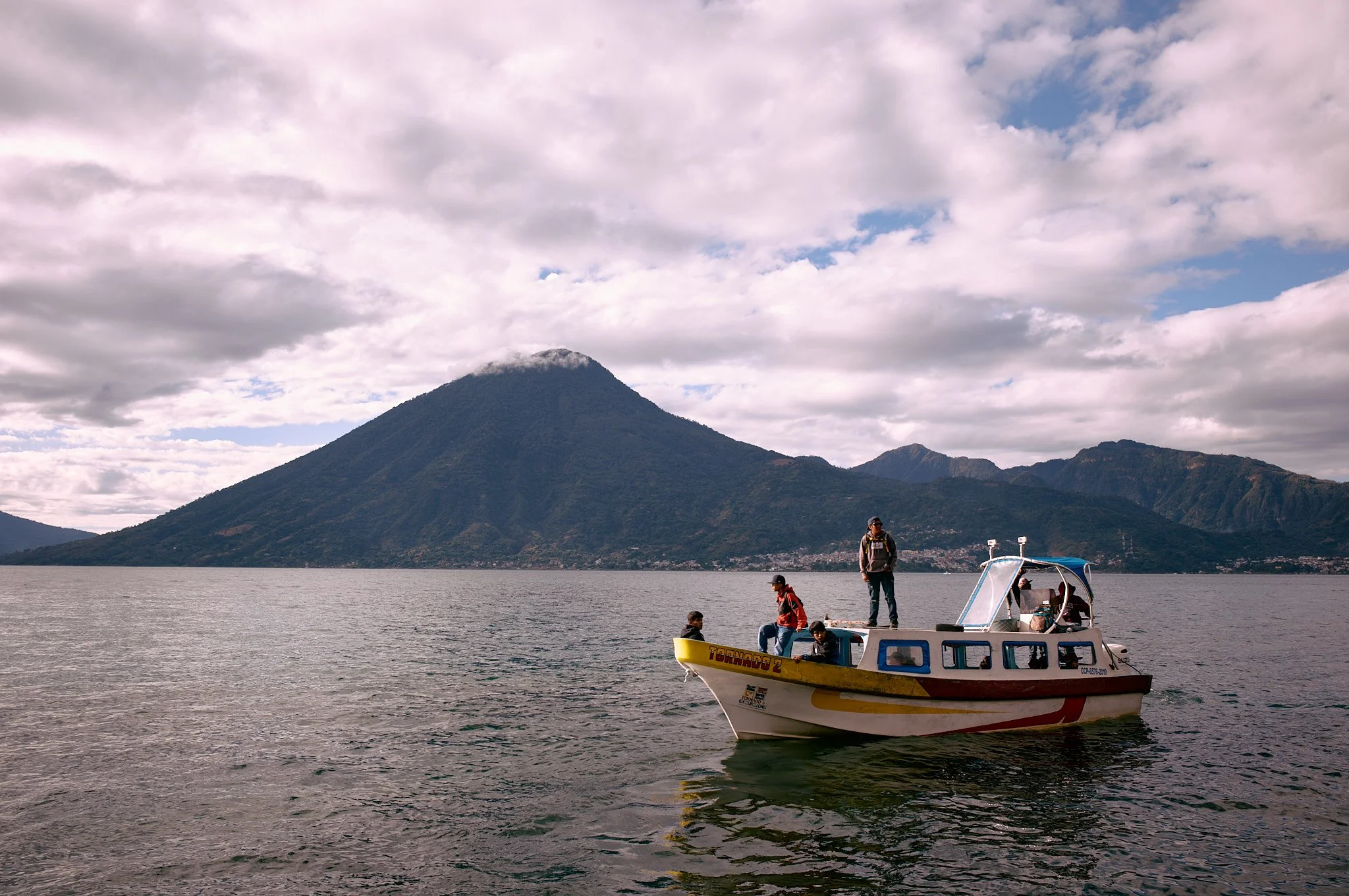 A group of people on a small white and yellow boat with a mountain in the background, on a body of water under cloudy skies.