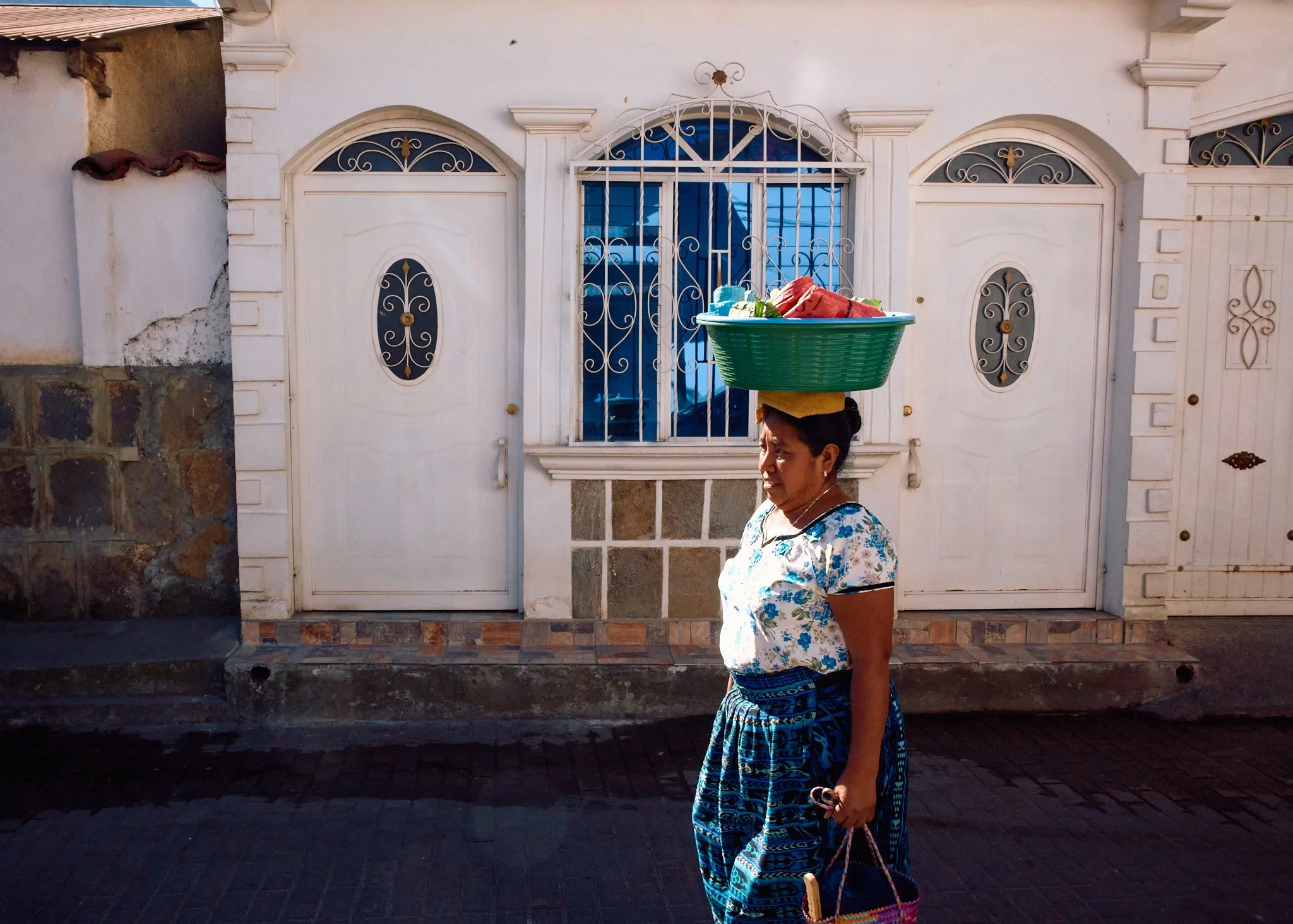 A woman wearing traditional clothing, carrying a green plastic basket full of items on her head, walking past a white house with ornate window and door ironwork.