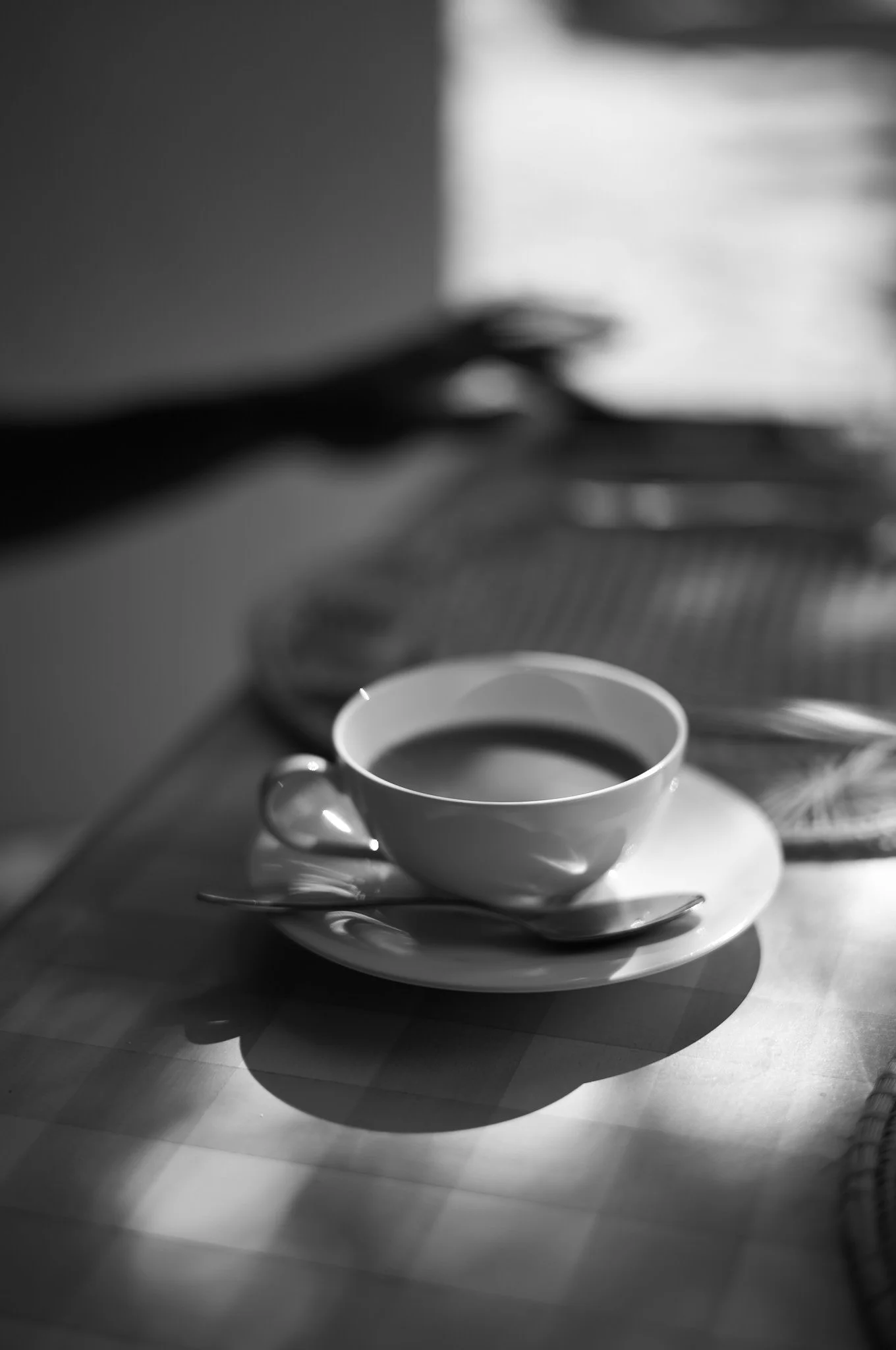 A black and white photo of a cup of coffee on a saucer with a small spoon resting on it, placed on a checkered table.