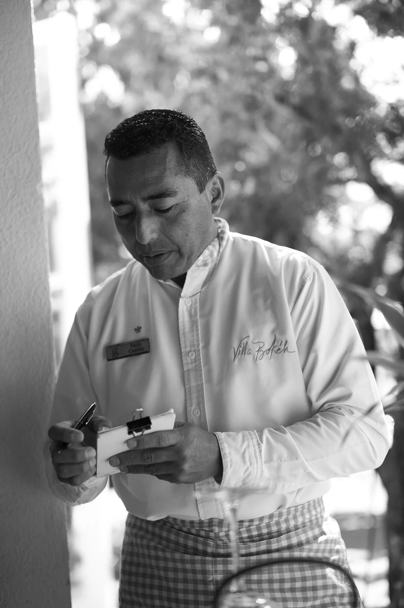 A man wearing a chef's coat and checkered apron, writing on a notepad, standing outdoors with blurred trees in the background.