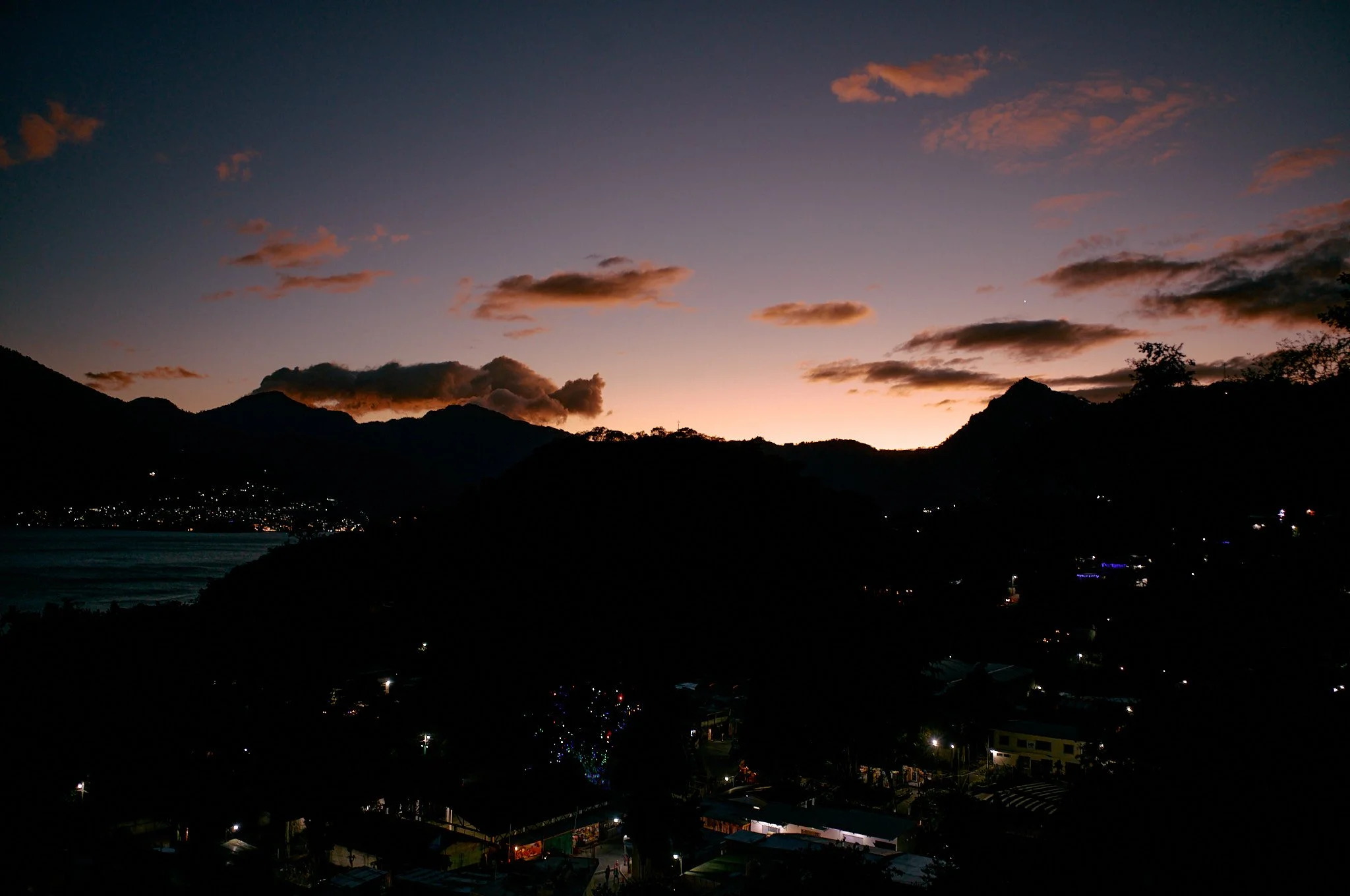 Sunset over mountainous landscape with clouds and a town with lights in the foreground.