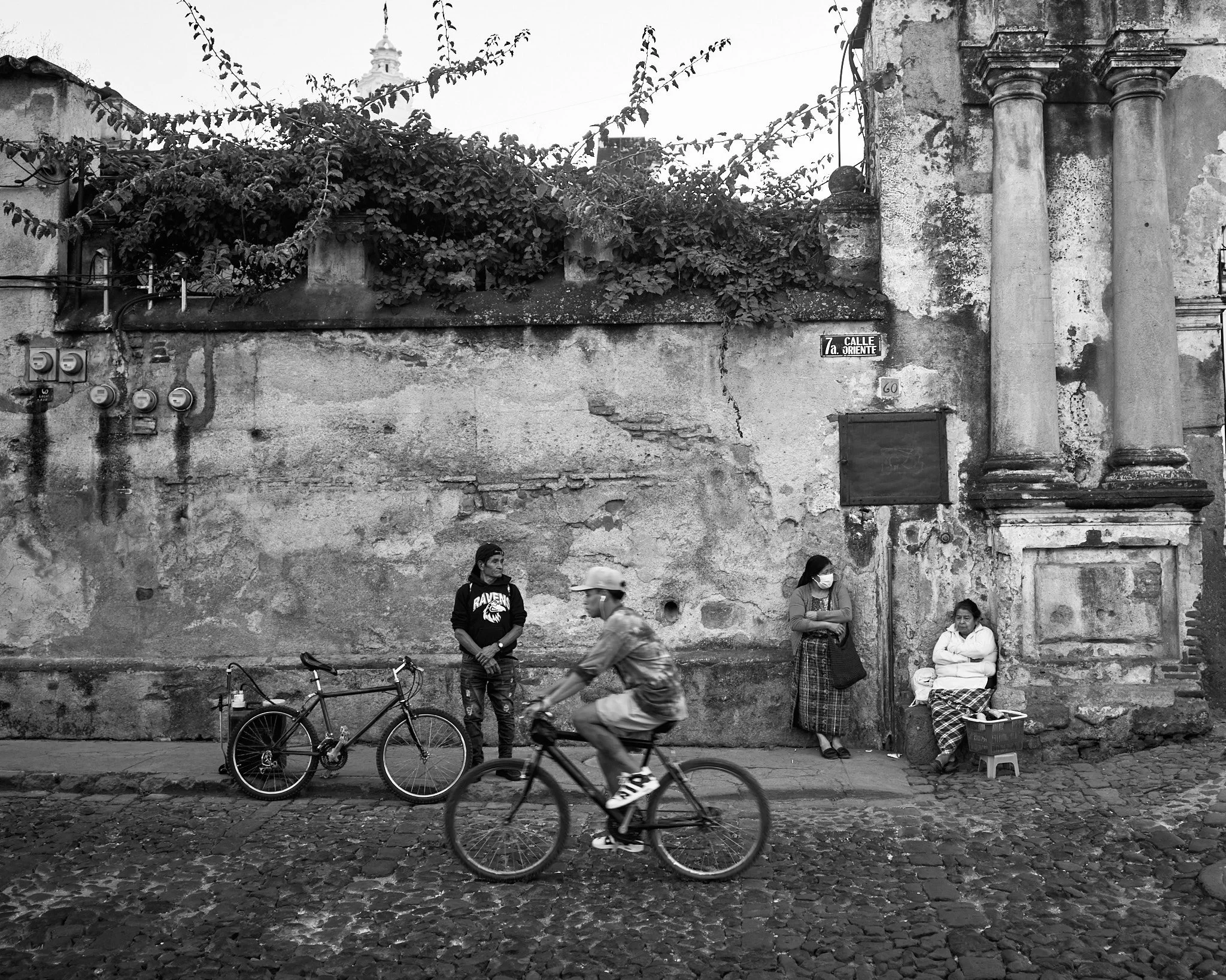 Street scene with three people standing and one riding a bicycle, on a cobblestone road in front of a weathered wall. One woman is sitting on a small stool, another woman is leaning against the wall, and a third person is riding a bike past them.