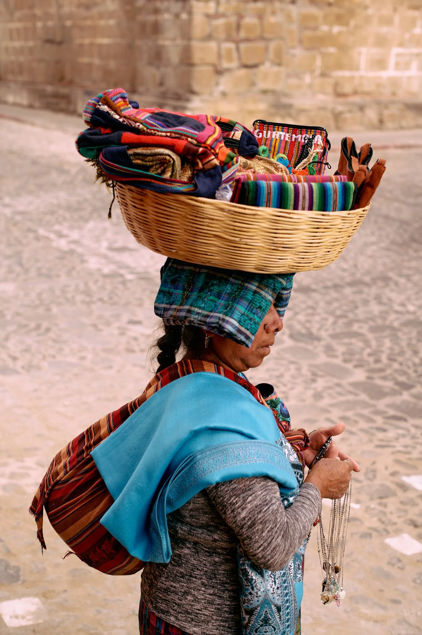 A woman with traditional colorful clothing and a head covering carrying a basket on her head filled with fabric and accessories, holding jewelry in her hands, standing on a cobblestone street with a brick wall in the background.