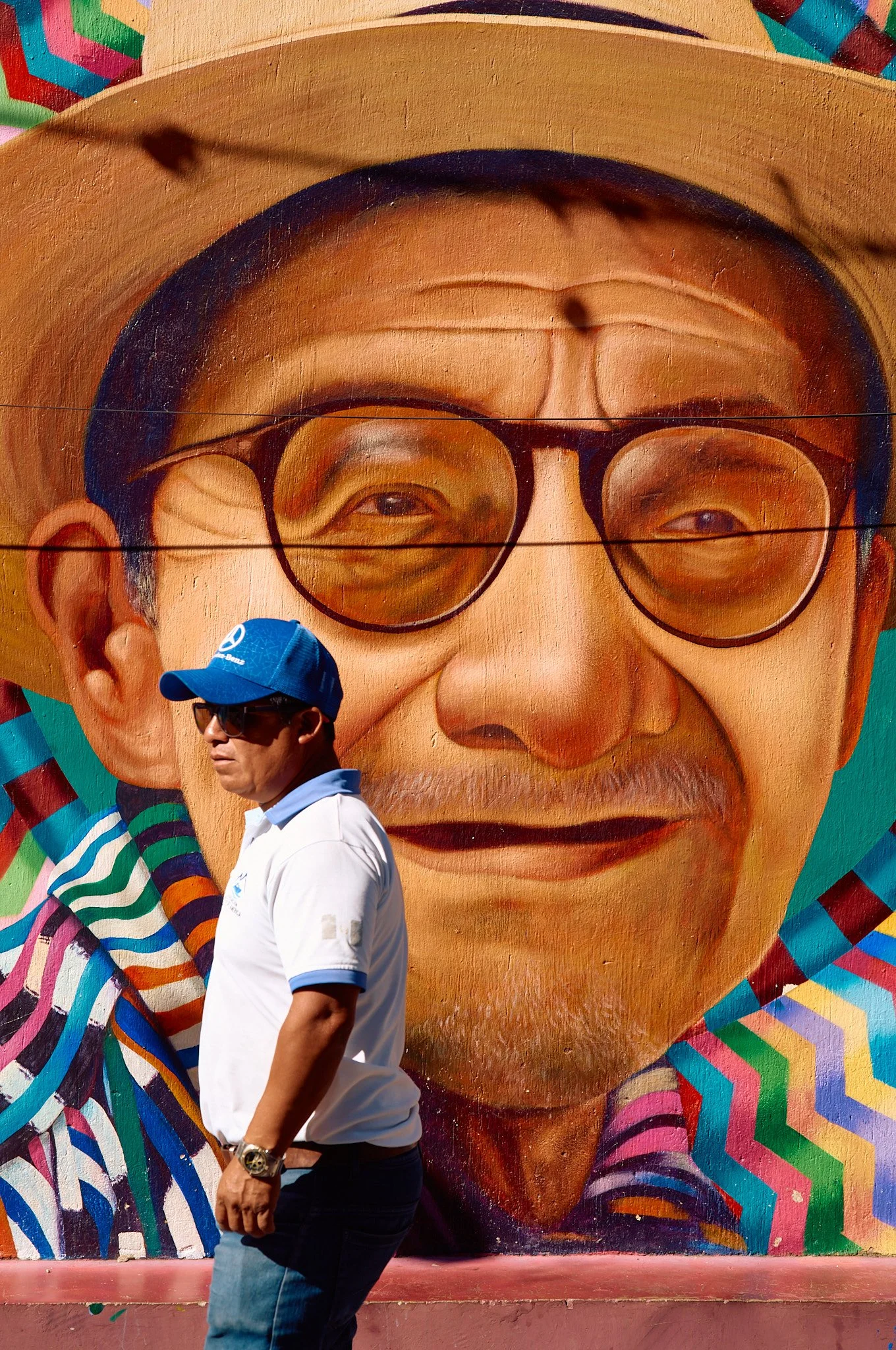 A man in sunglasses, a white polo shirt, and a blue cap is walking past a colorful mural of an elderly man with glasses, a hat, and a mustache.