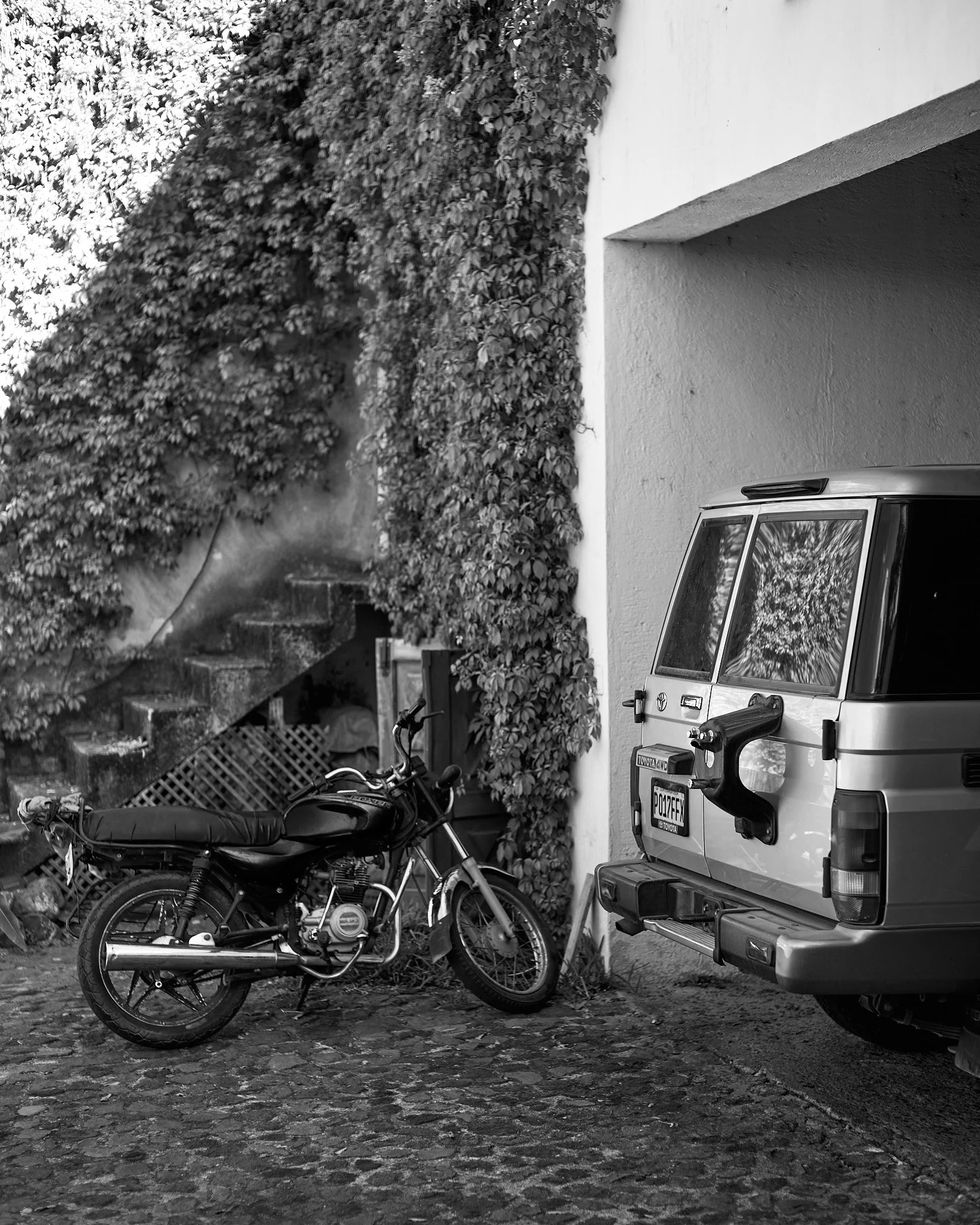 A black motorcycle parked next to a silver van under a shaded area with a wall and stairs covered in ivy in the background.
