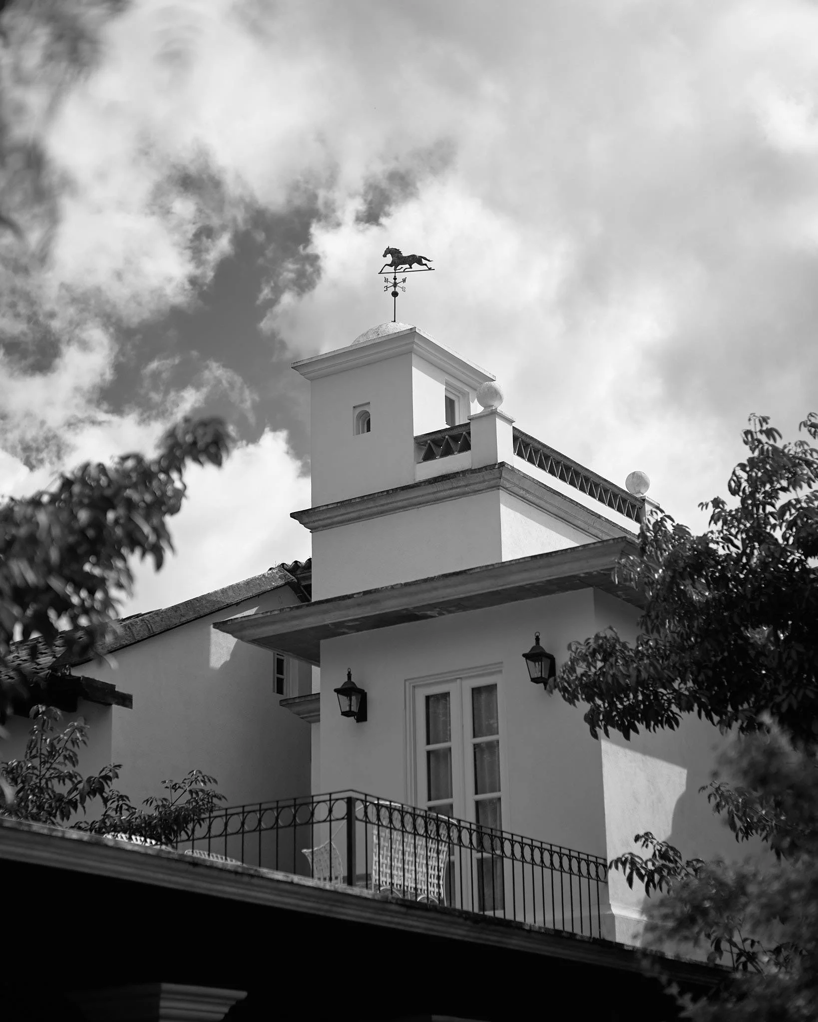 View of a white house with a rooftop weather vane in the shape of a horse, visible against a cloudy sky, surrounded by trees.