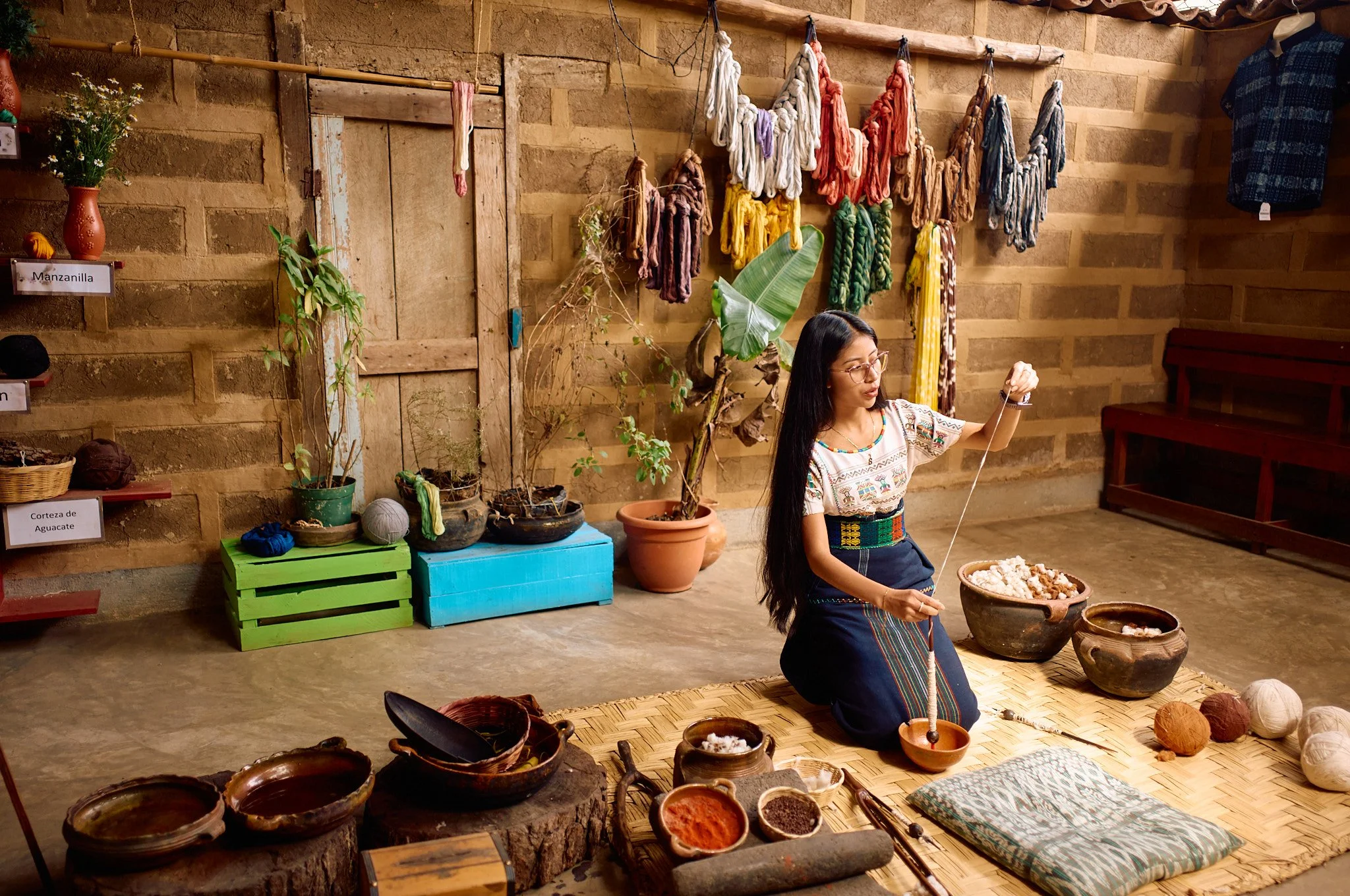 A woman kneeling on a woven mat, using a drop spindle to spin yarn. She is surrounded by bowls of natural materials, with colorful skeins of dyed yarn hanging on the wall behind her. There are potted plants and woven baskets in the background.