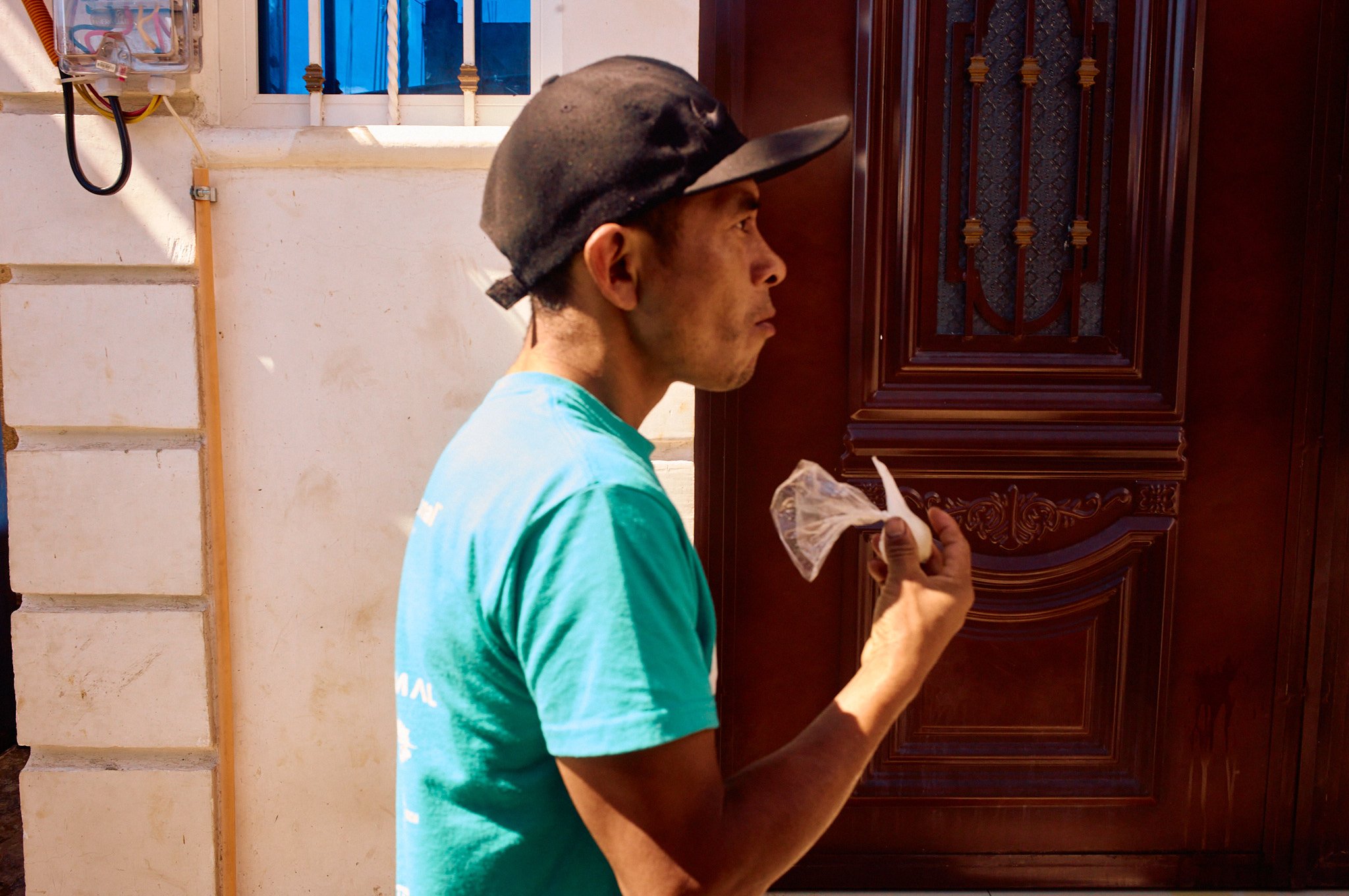A man with a cap holding a plastic bag in front of a wooden door outside a house.