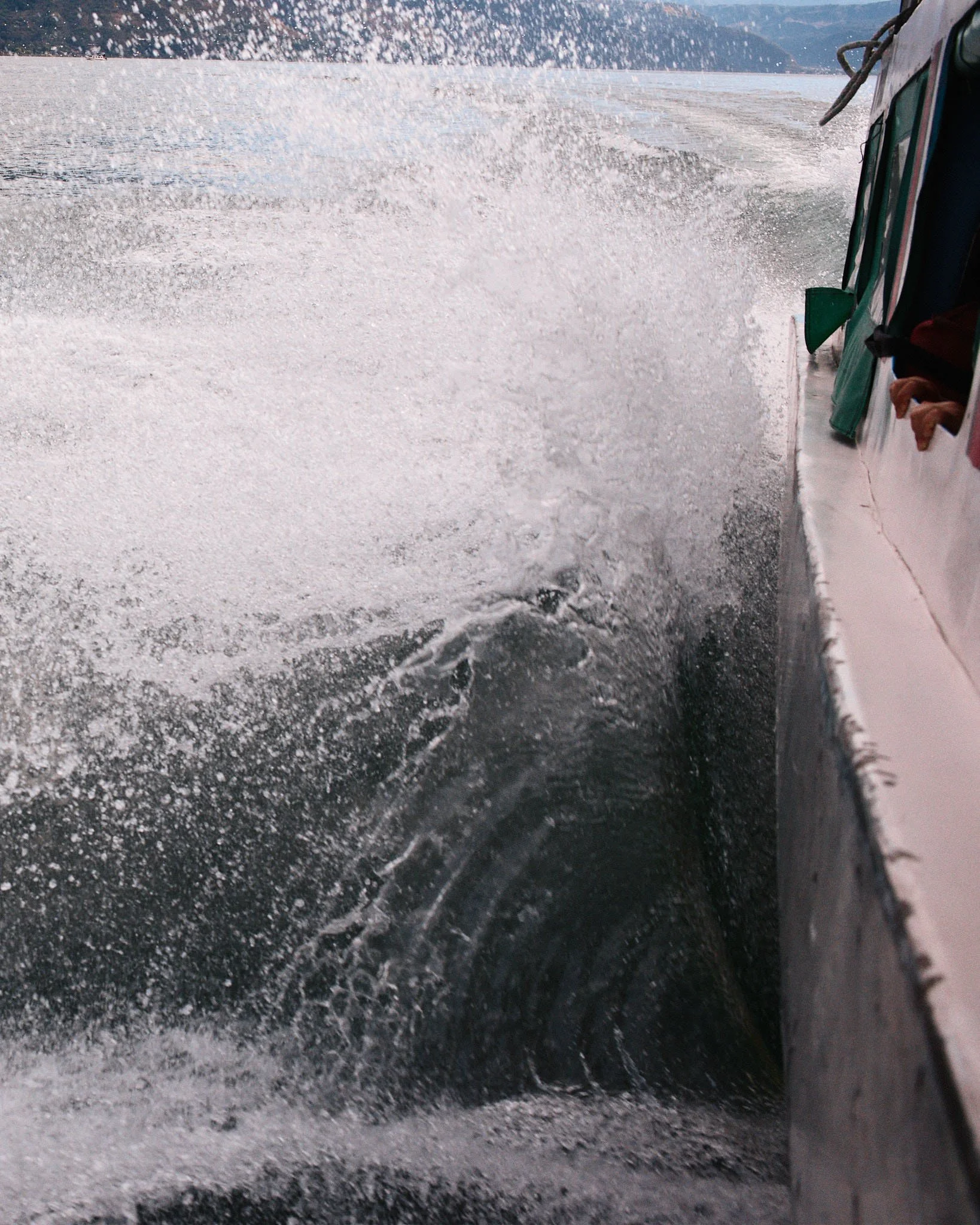 View from a boat showing the wake and spray in the water, with a mountainous landscape in the background.
