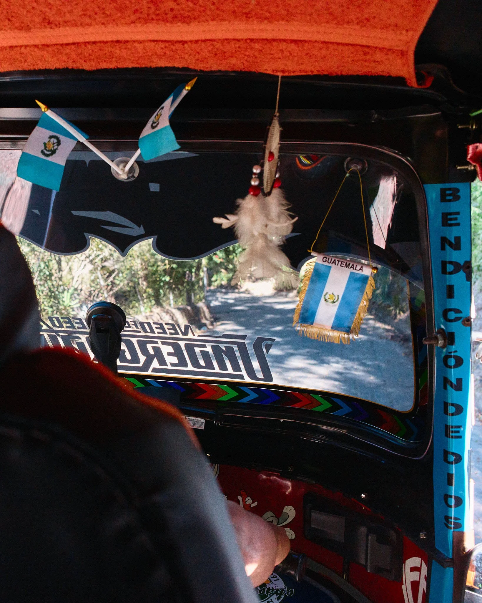 Inside a tuk-tuk decorated with Guatemalan flags, a hanging feather ornament, and a small Guatemala flag with a yellow fringe. The view shows a rural road through the front windshield.