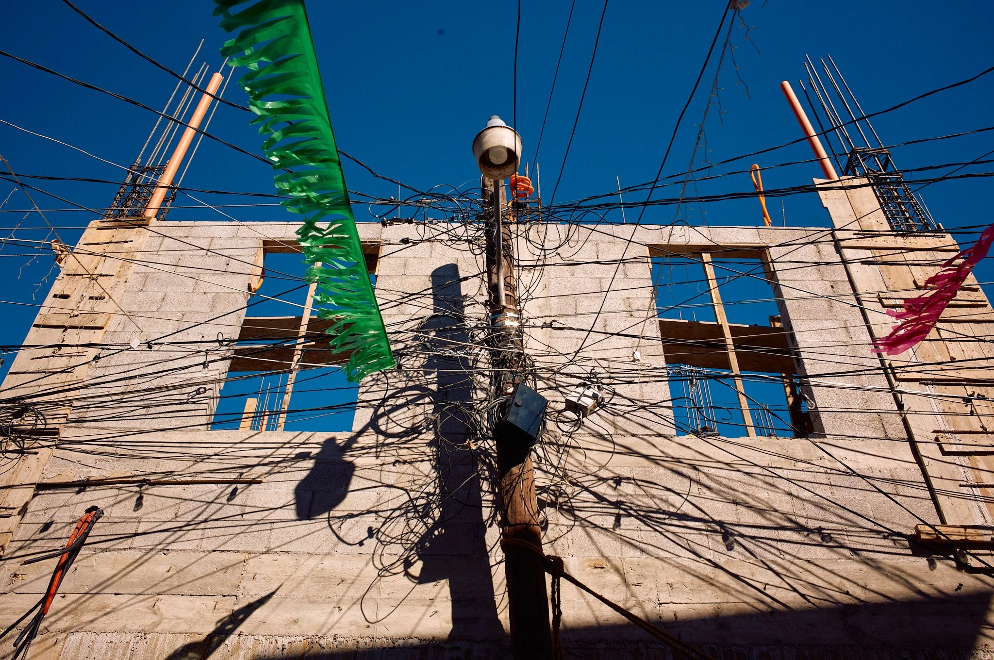 A building under construction with exposed brick walls, two window openings, and electrical wires tangled around a utility pole in front of it, under a clear blue sky.