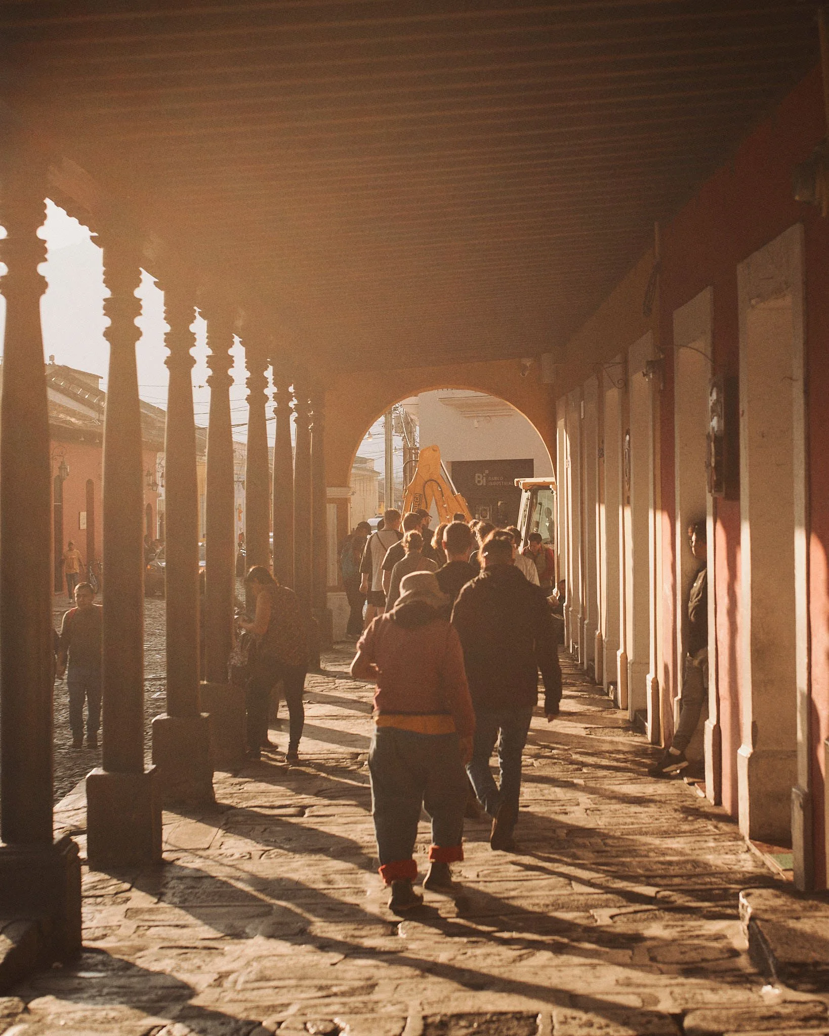 People walking along a covered walkway with columns, sunlight casting shadows, and an excavator visible in the background.