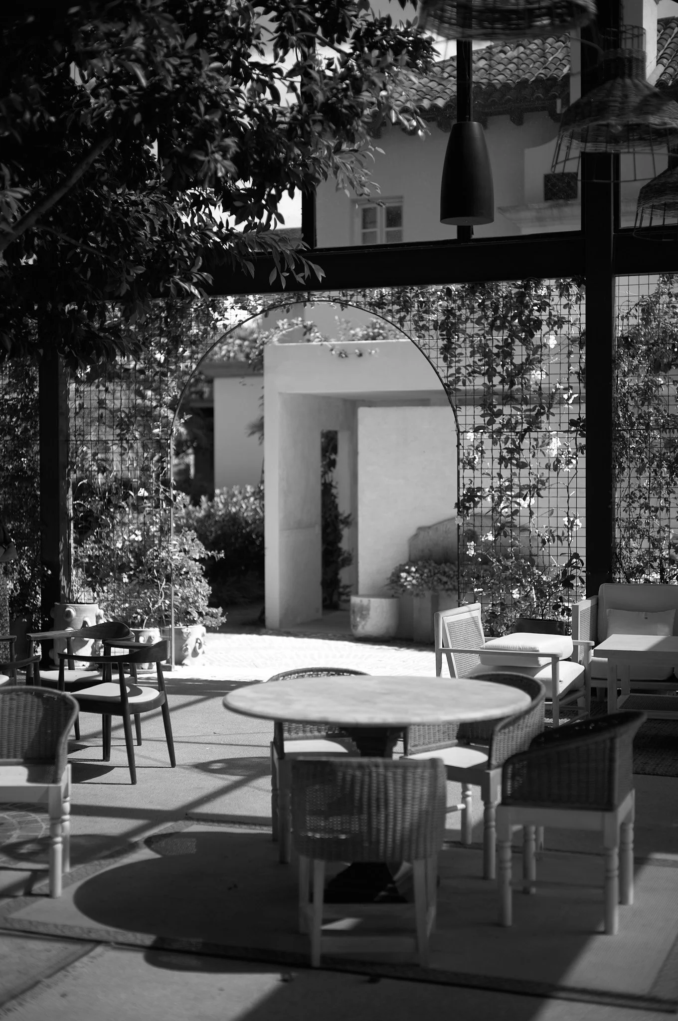 A black and white photo of a modern outdoor patio with round and rectangular chairs, a circular table, a rug, hanging pendant lights, and an archway leading to a garden with plants and a white wall.