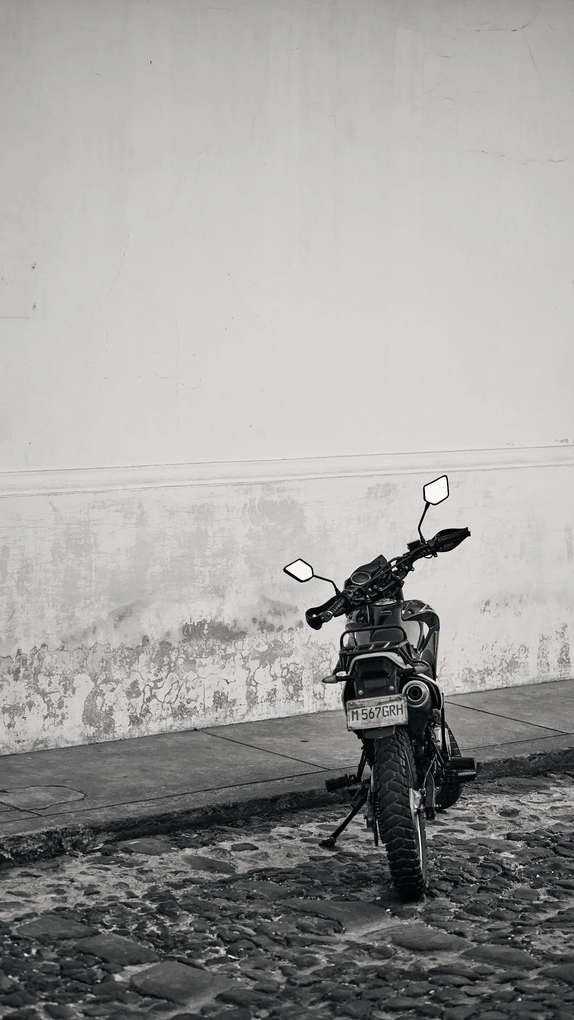 A black motorcycle parked on a cobblestone street next to a weathered white wall with peeling paint.
