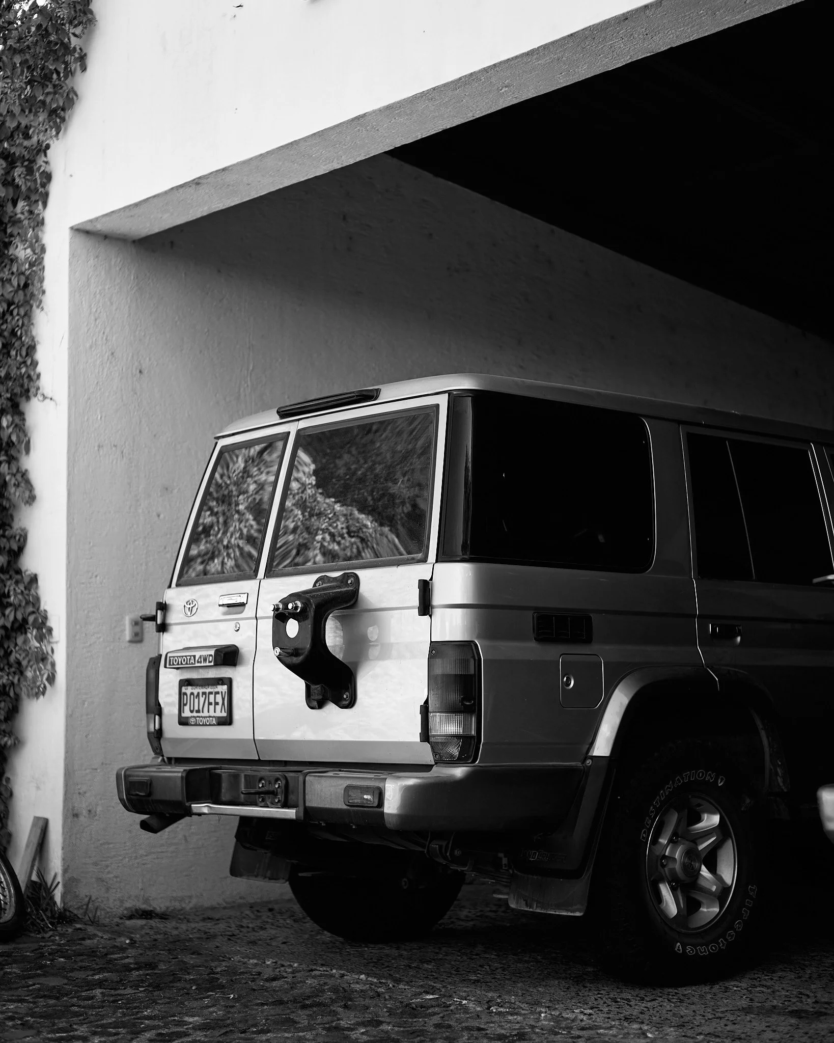 Black and white photo of a vintage Toyota 4WD SUV parked under a building's overhang, with plants visible on the left side.