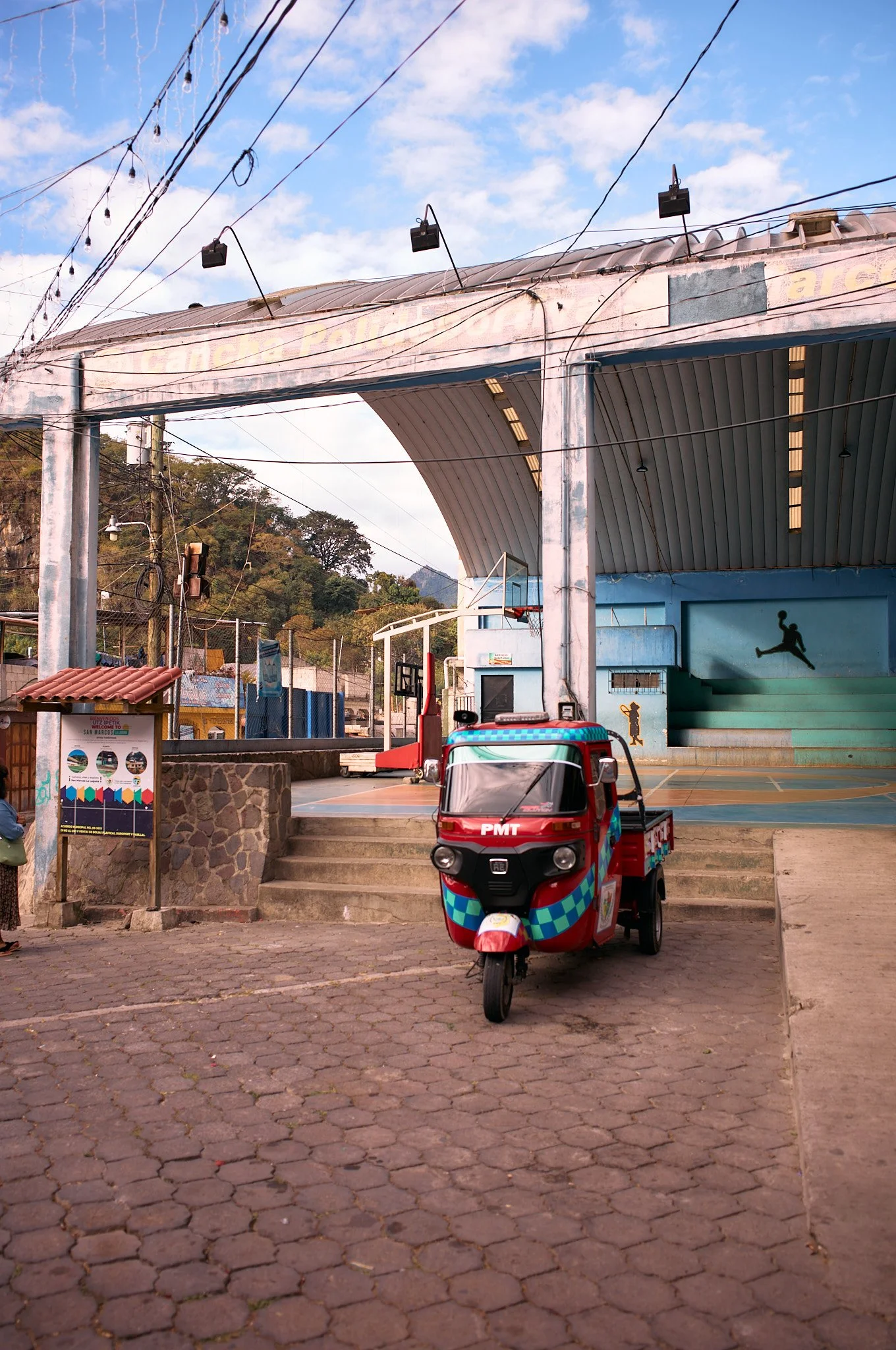 A small tuk-tuk parked outside a basketball court with painted steps, covered by a large, curved roof. The court has a basketball hoop and painted silhouettes of basketball players. There are string lights hanging overhead, and a mountain can be seen