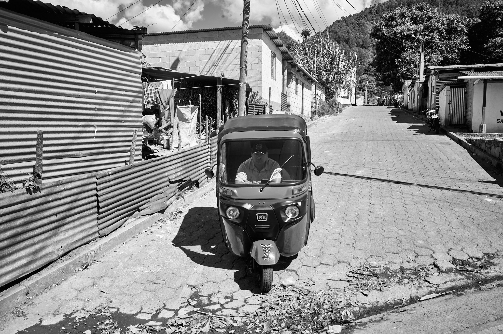 A man driving a three-wheeled motorcycle on a sloped paved road in a rural area with houses and trees on either side