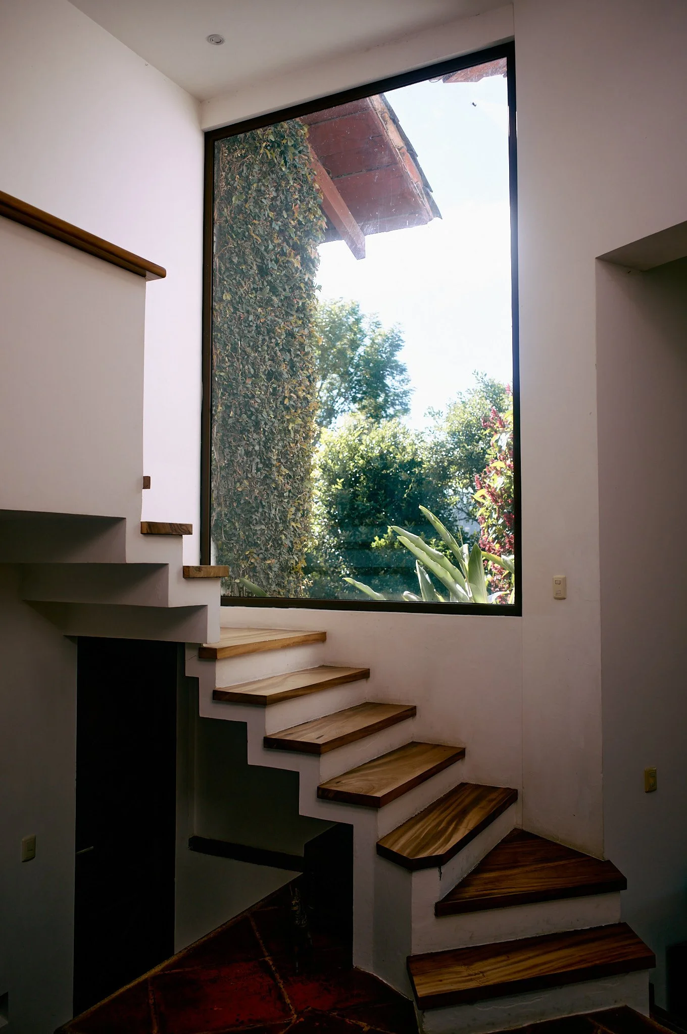 Interior of a house with wooden stairs leading up past a large window showing greenery outside.