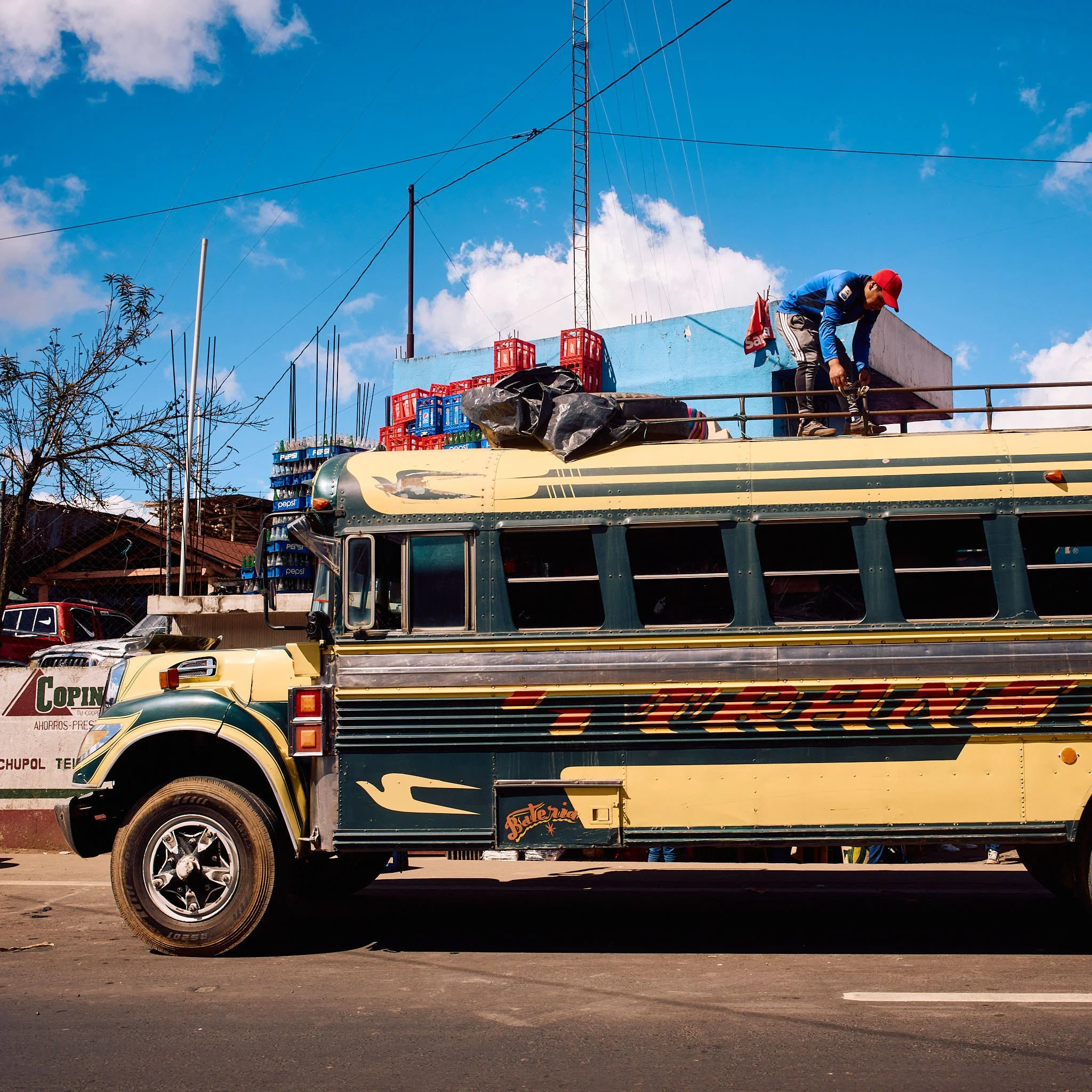A large bus with a man on top loading or unloading goods, parked on the street with blue sky and clouds in the background.