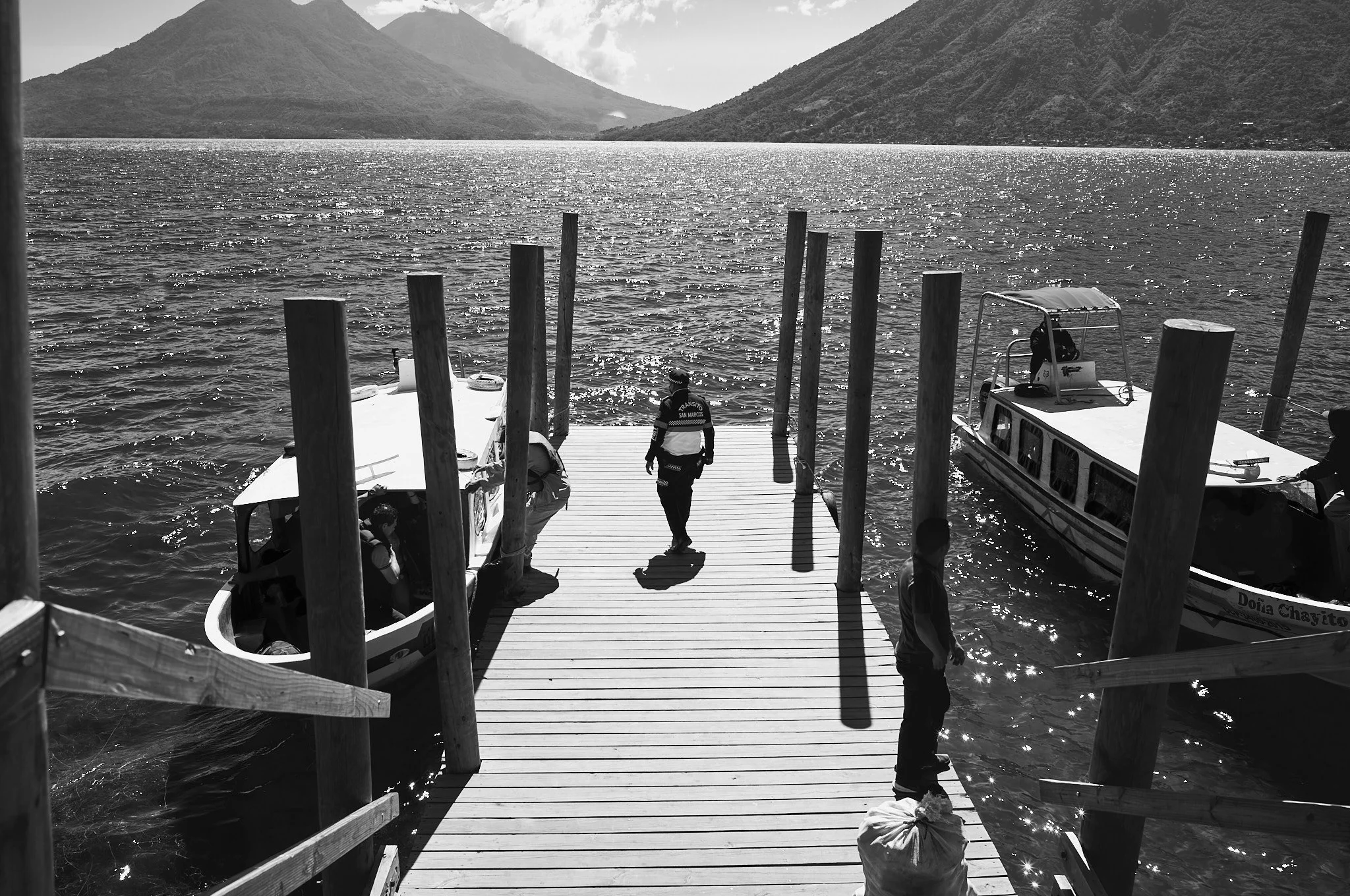 A wooden dock extending into a lake, with two boats on either side, and mountains in the background. Several people are on or around the dock, some sitting in boats and others standing or walking.